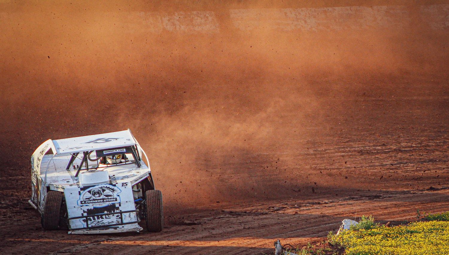 A dirt track racing car kicks up a massive cloud of red dust while speeding around a muddy oval circuit.