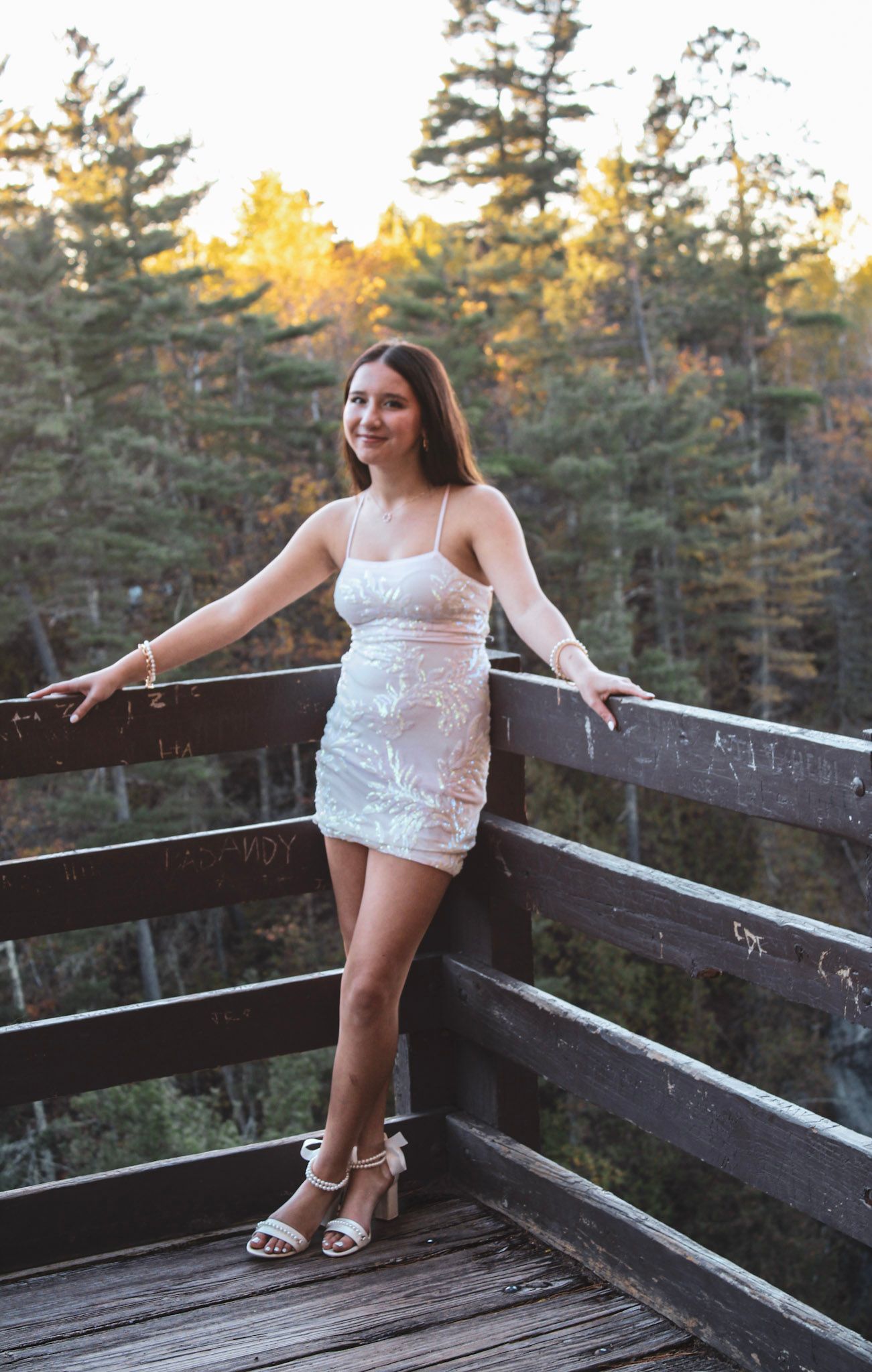 A person poses in white dress on wooden observation deck surrounded by pine forest.