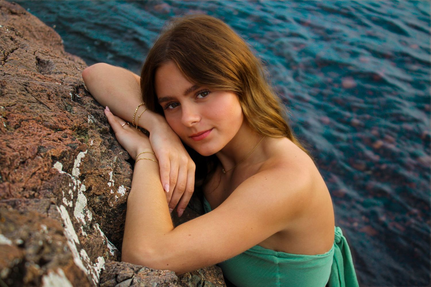 Young woman in green dress poses gracefully against rocky coastal backdrop with blue water behind her.