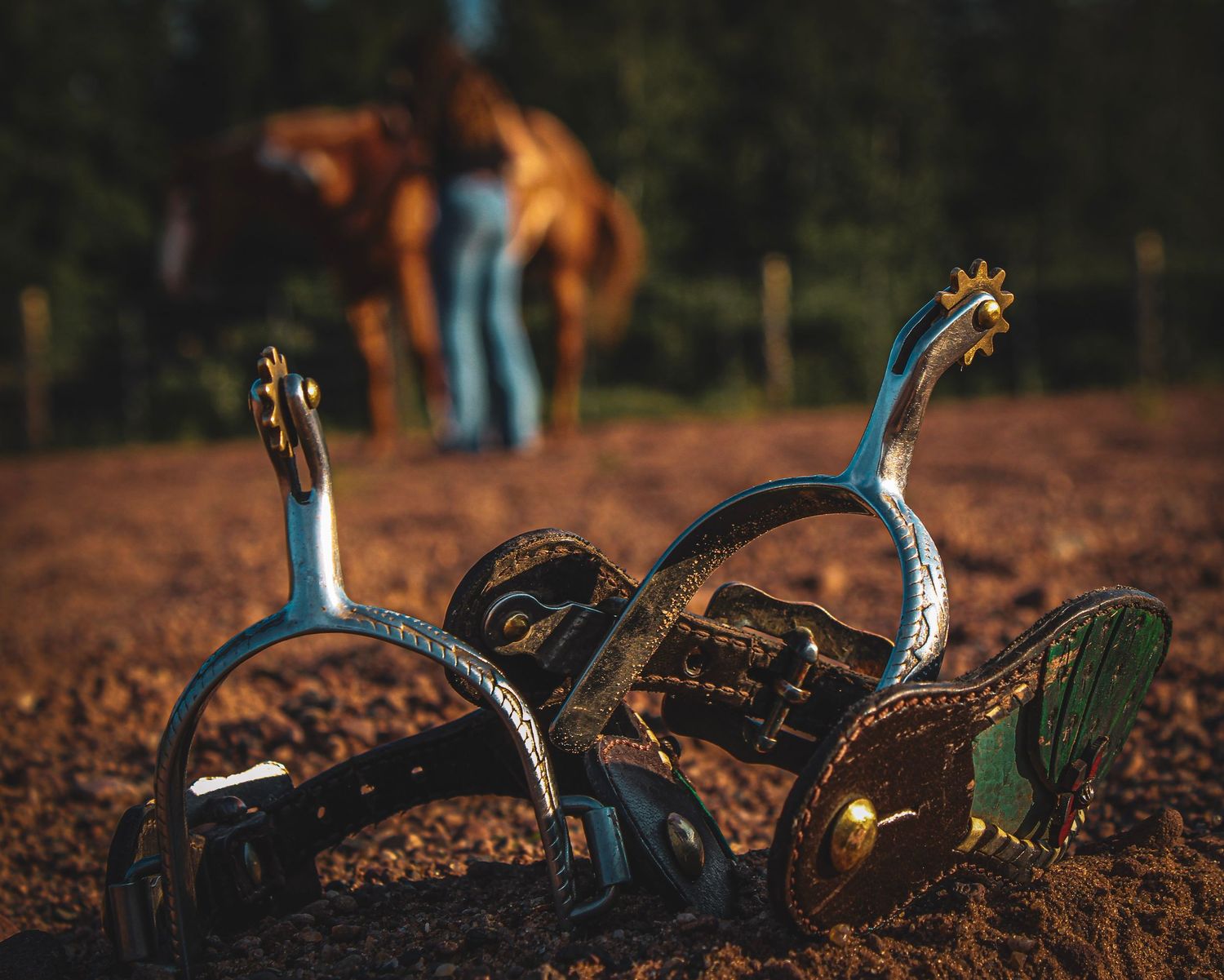 Decorative Western spurs with gold accents rest on dirt ground, with a horse and rider blurred in the background.