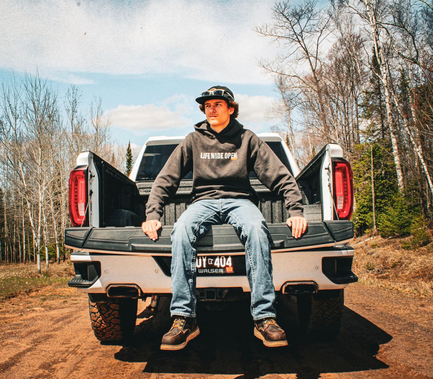Young man in hoodie and cap sits on tailgate of pickup truck on dirt road surrounded by trees.