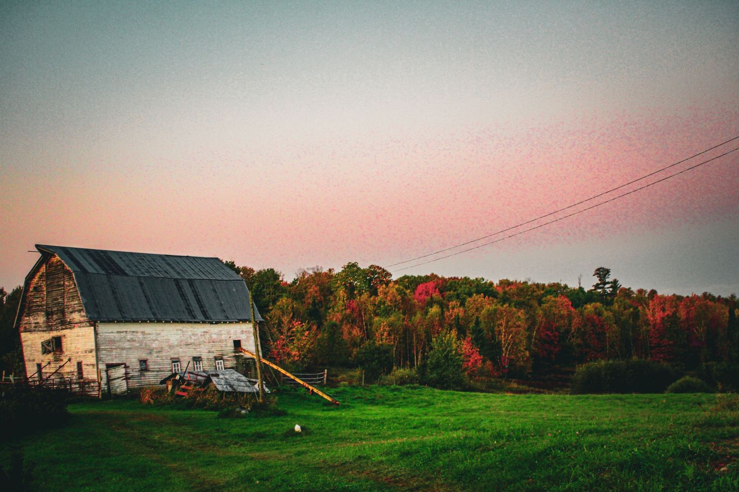 A scenic panoramic view of rural farmland with autumn foliage, barns, and power lines against a pink sunset sky.