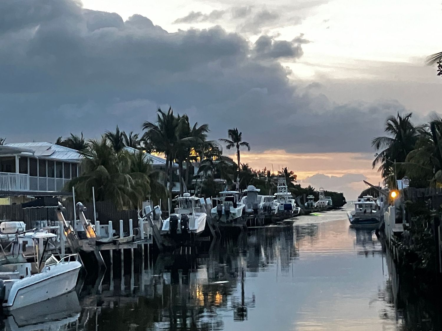 Dramatic sunset over waterfront homes and docked boats along a residential canal with palm trees and cloudy skies.