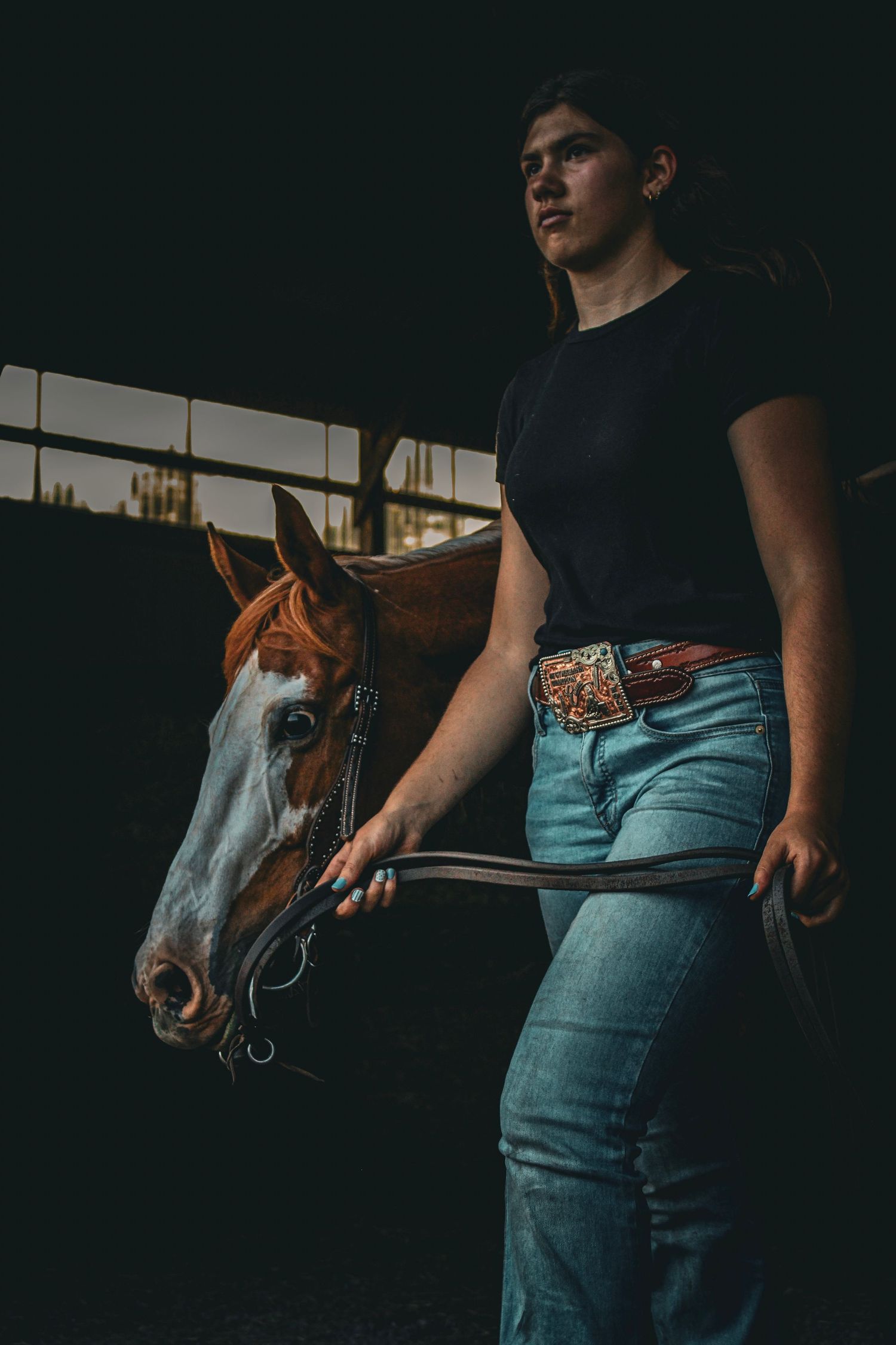 Young woman in jeans and black tee leads a chestnut horse by the reins inside a dark stable.