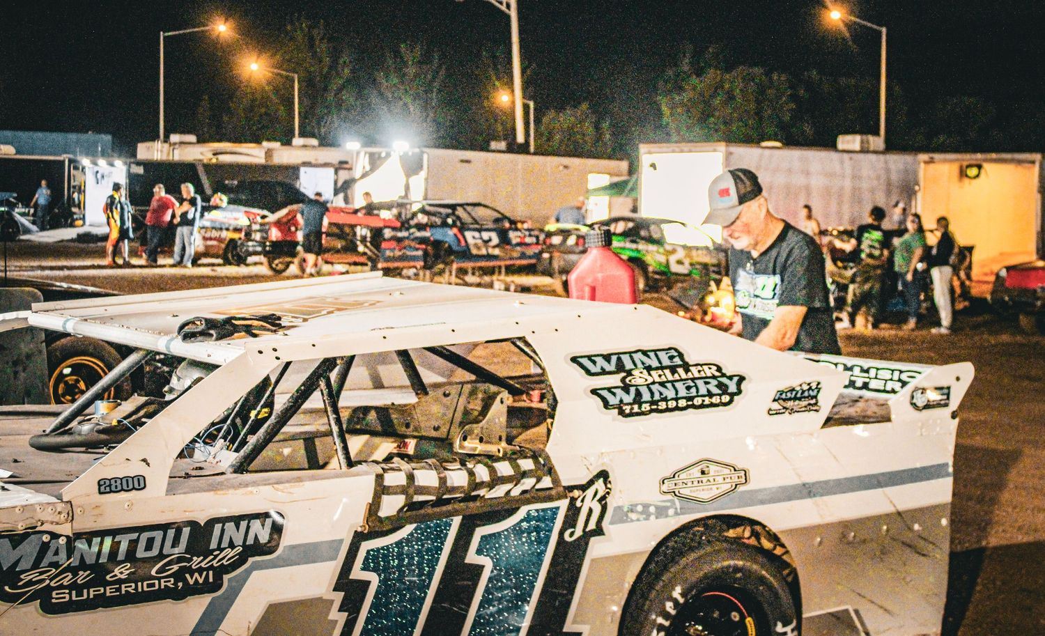 Super stock, stock cars line up under bright lights at a dirt track racing event during nighttime competition.