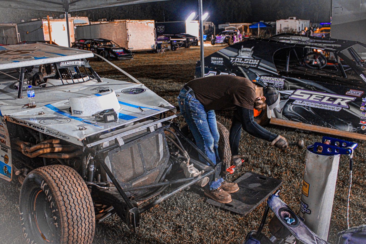 Racing vehicles lined up for maintenance with crews working in evening lighting.