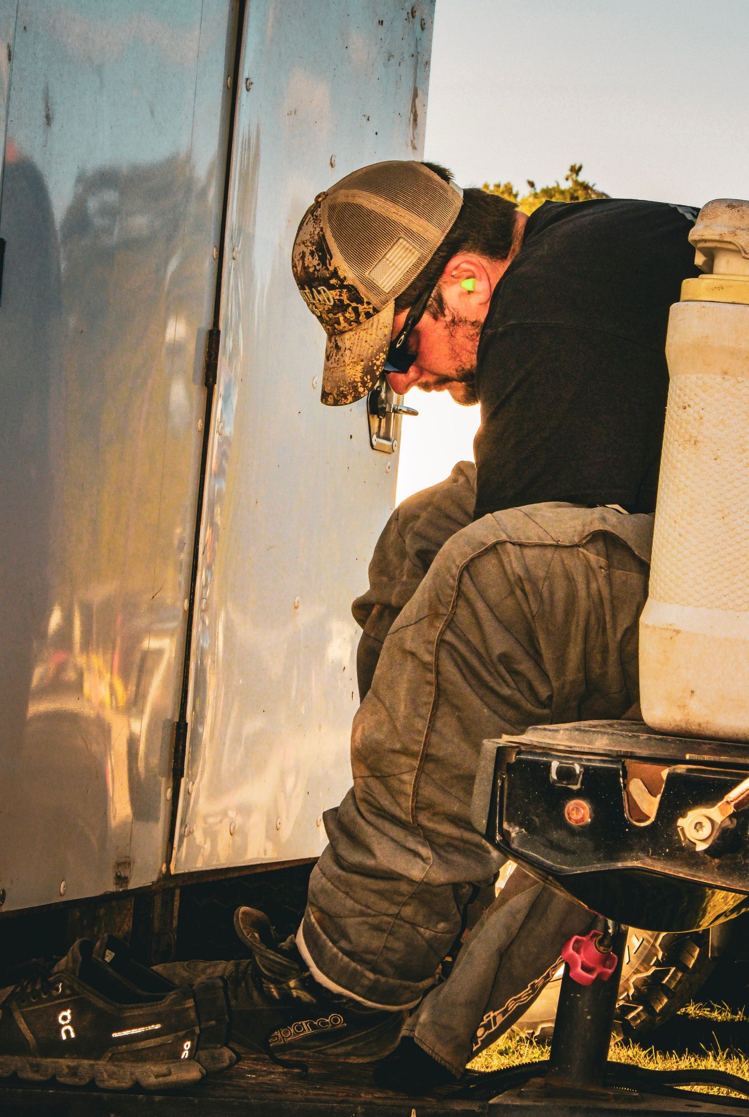 Crew member in camo hat working on race car during golden hour sunset.