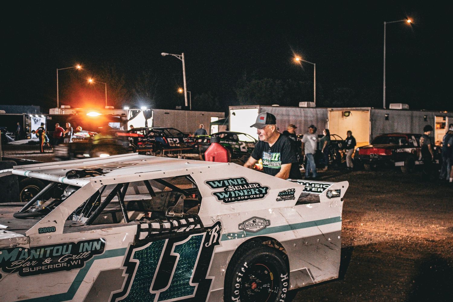 A white race car with number 111 sits at a dirt track racing event at night with spectators and lights in background.