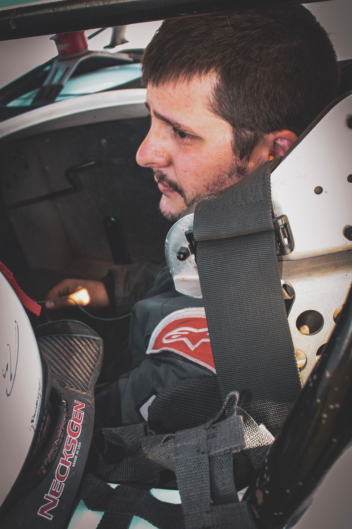 Race car driver in safety gear sits focused in cockpit wearing racing harness and protective equipment.