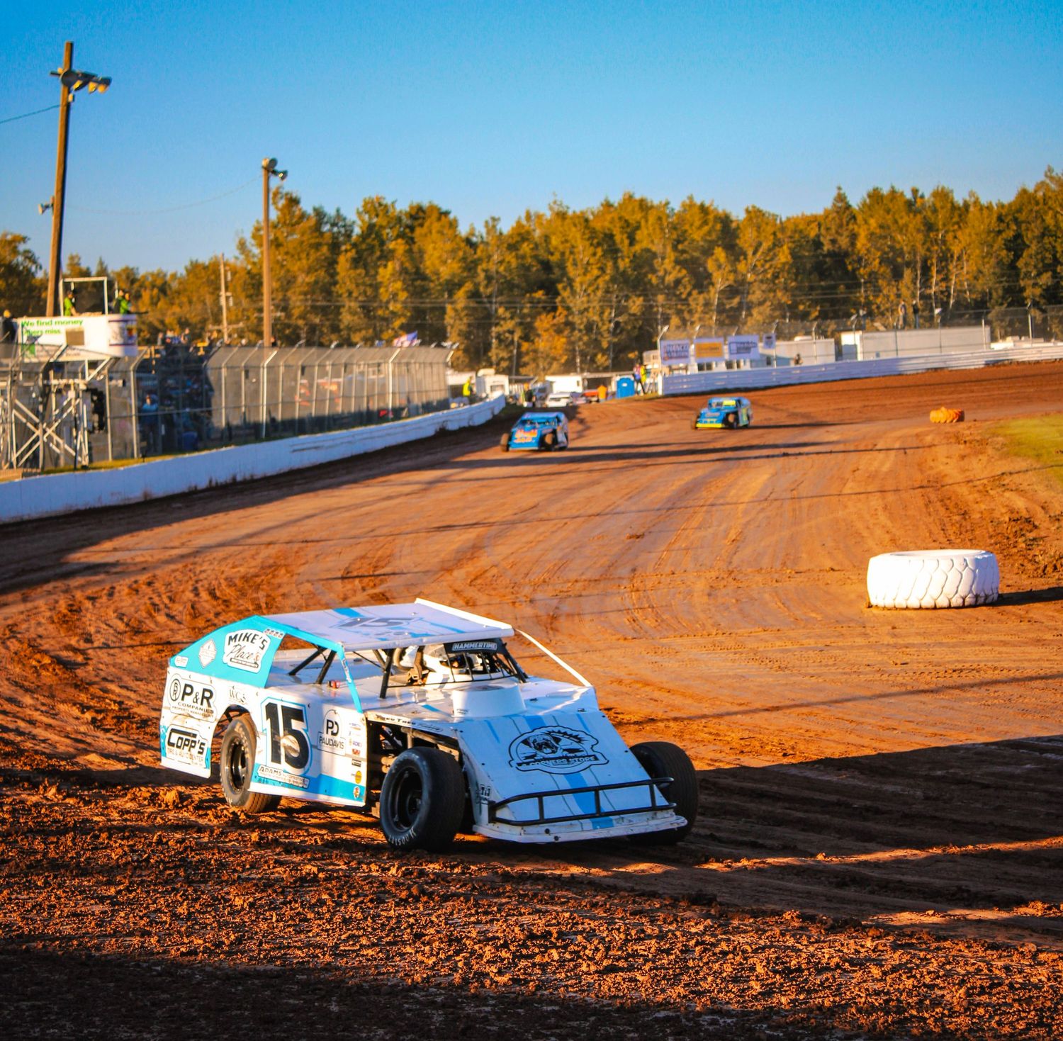 Dirt track racing cars competing on an oval circuit with autumn trees in the background.