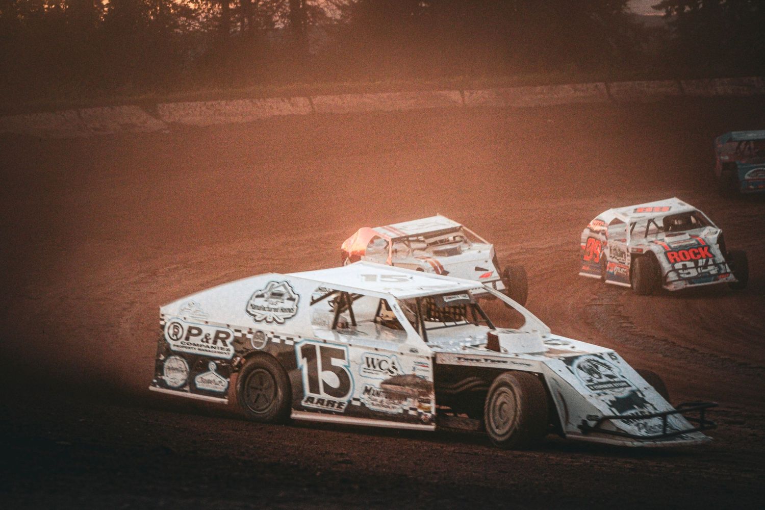 Dirt track racing cars competing on a dusty oval circuit at sunset, with car number 15 leading the pack.