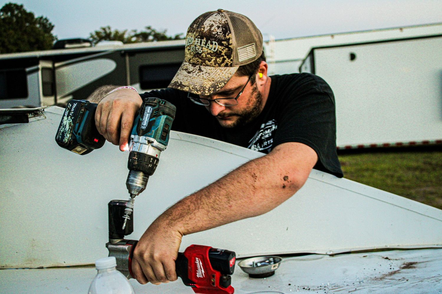 Mechanic in camo hat uses power drill while working on race car exterior.
