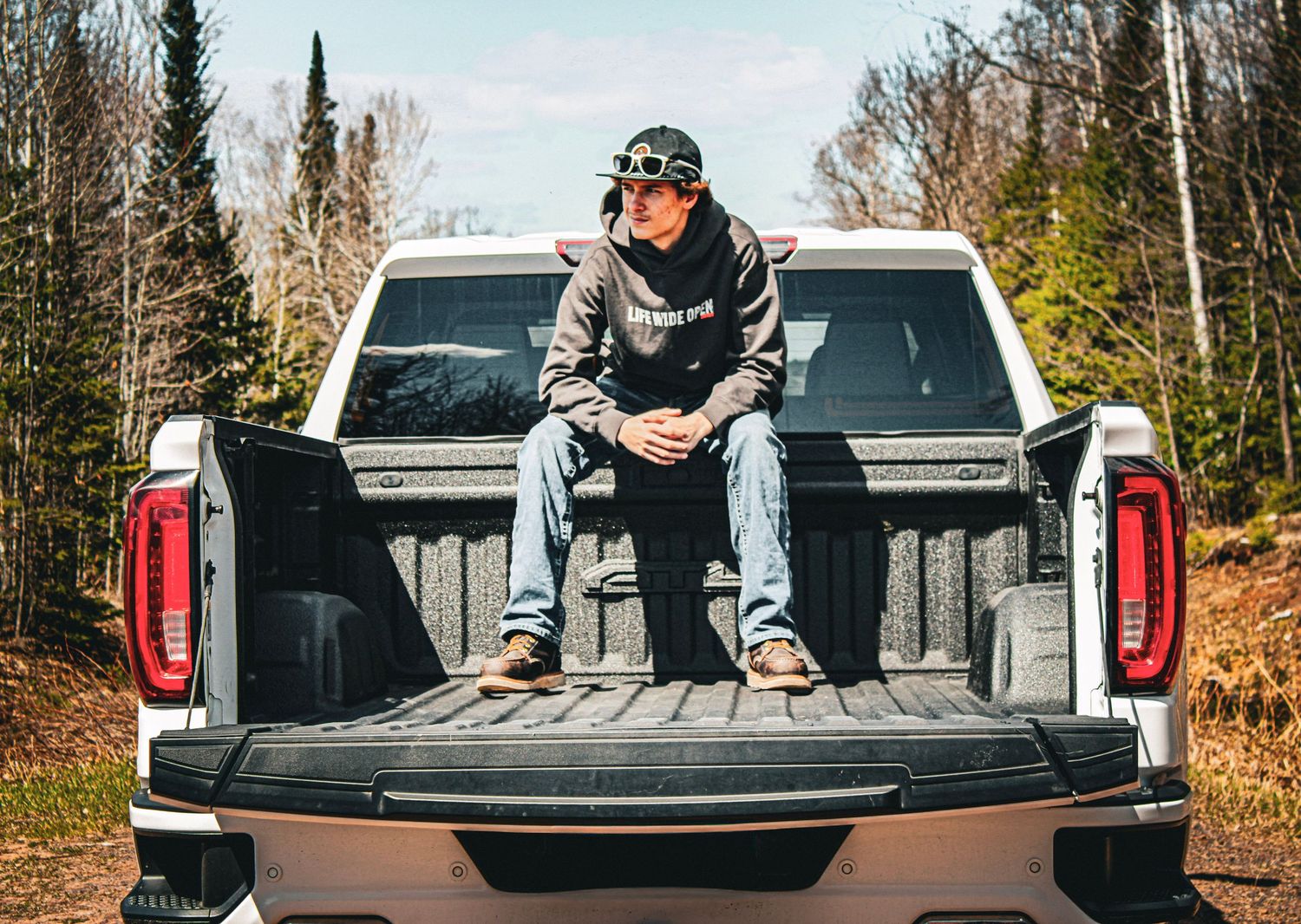 Young man in hoodie and cap sitting on the tailgate of a white pickup truck on a forest road.