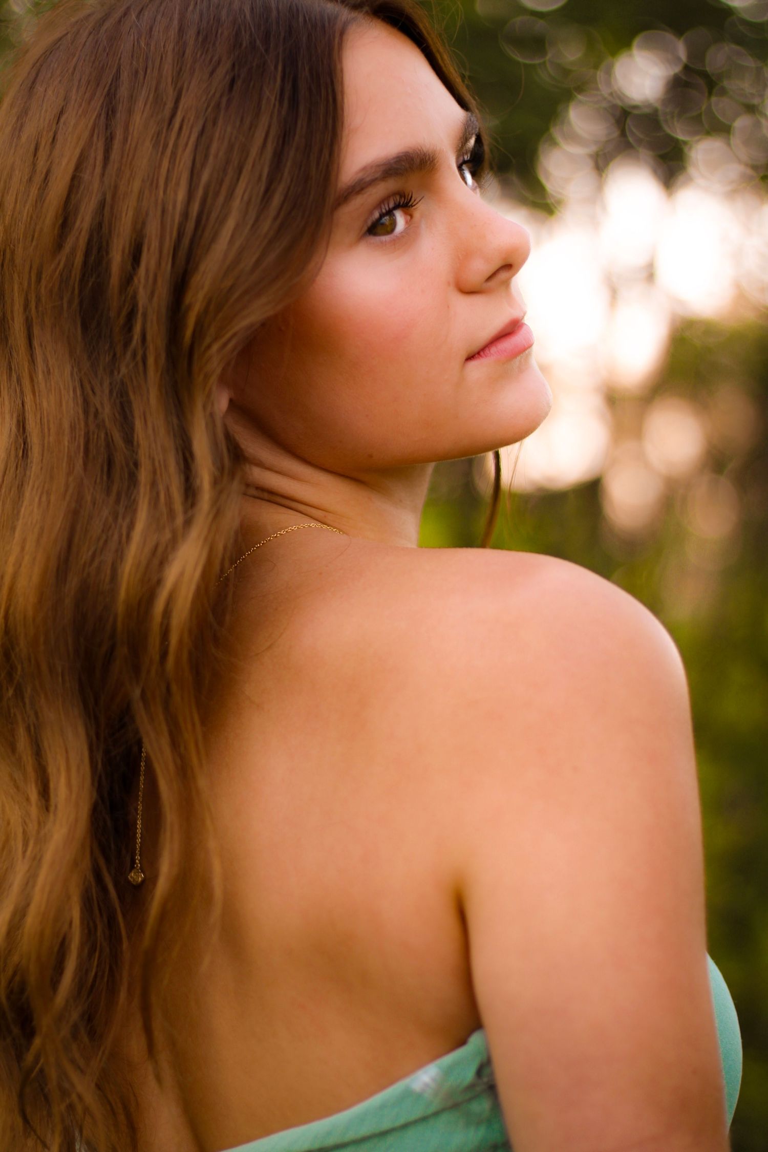Young woman with long brown hair in a strapless green dress, glancing over her shoulder outdoors.