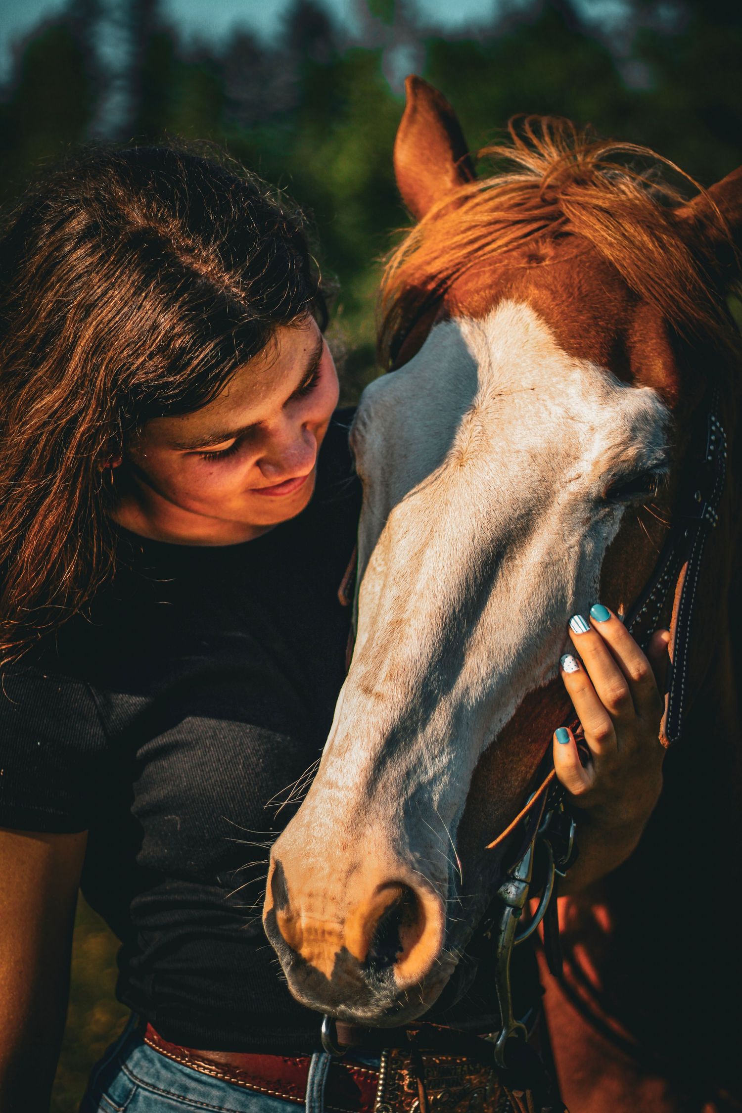 Young woman with long dark hair tenderly rests her forehead against a chestnut horse with a white blaze in golden light.