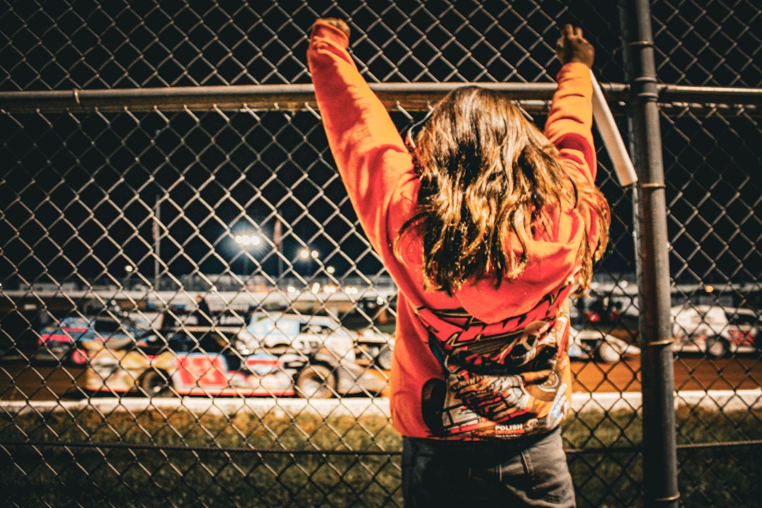 Person in orange jacket watching racing action through chain link fence at motorsports event.