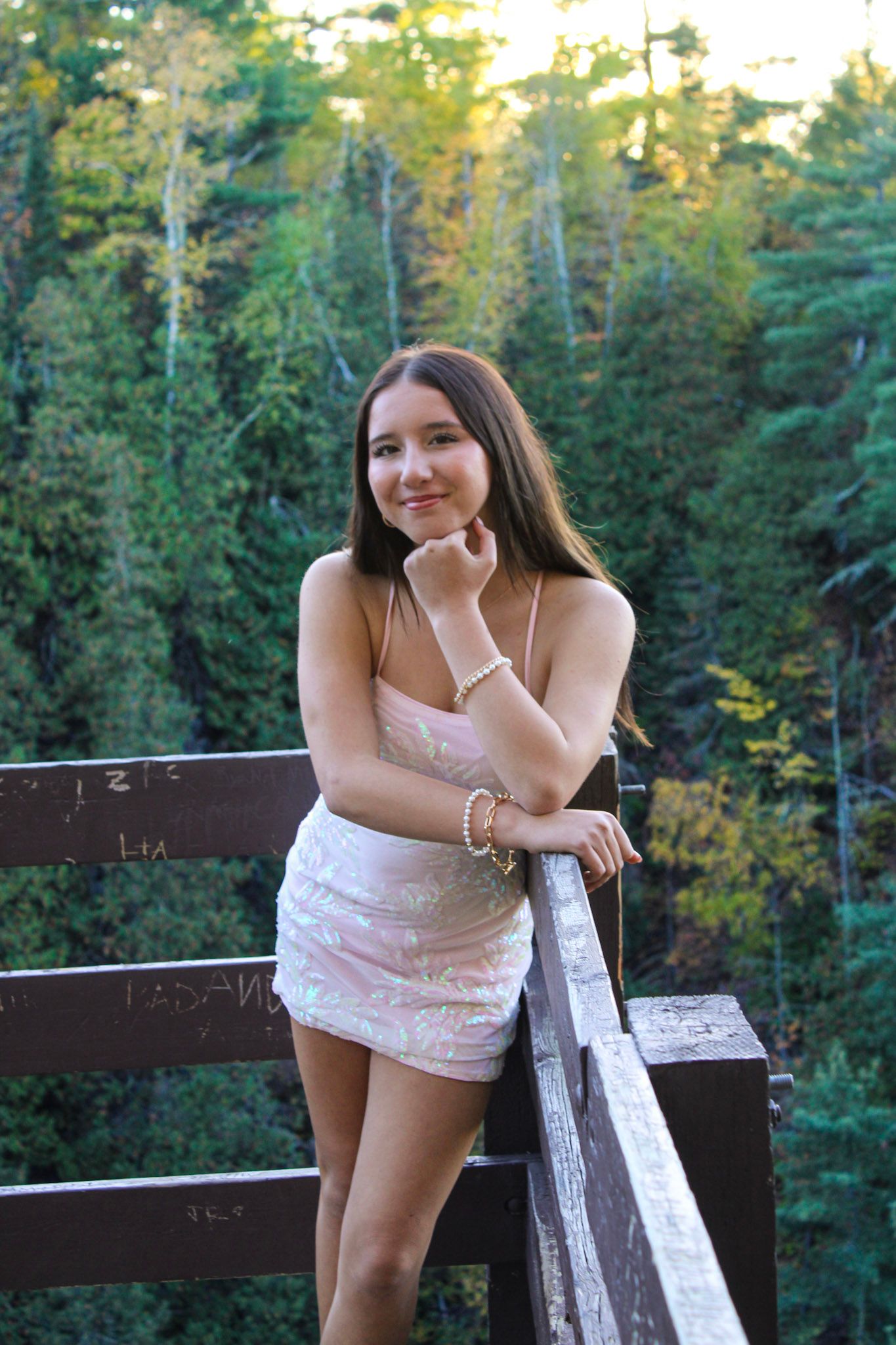 A person in white dress leans against wooden railing with forest scenery behind.
