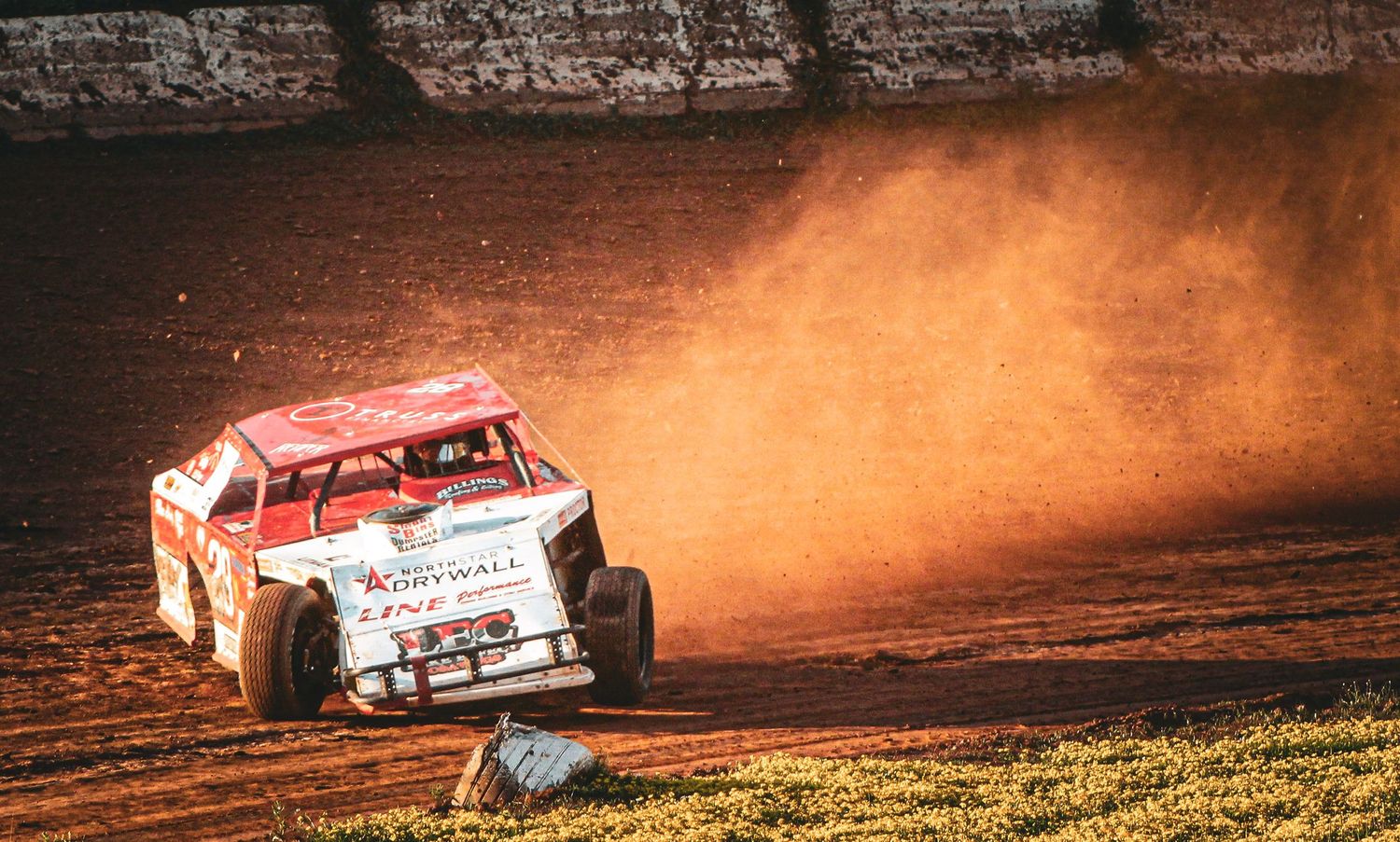 A dirt track racing car kicks up a cloud of dust while navigating a turn on a red clay oval racing circuit.
