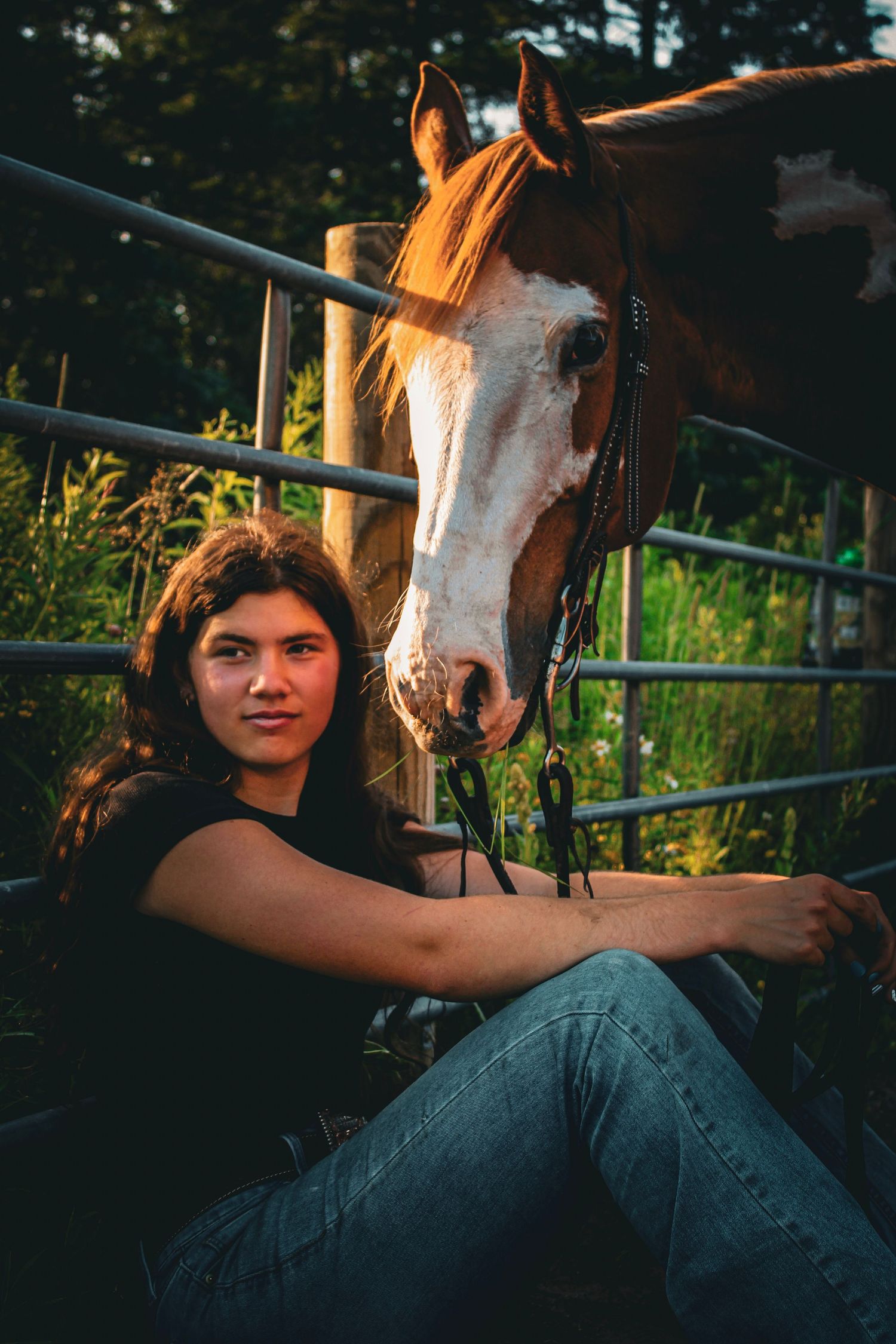 Young woman in black top and jeans sits against a metal fence beside a brown and white horse outdoors.