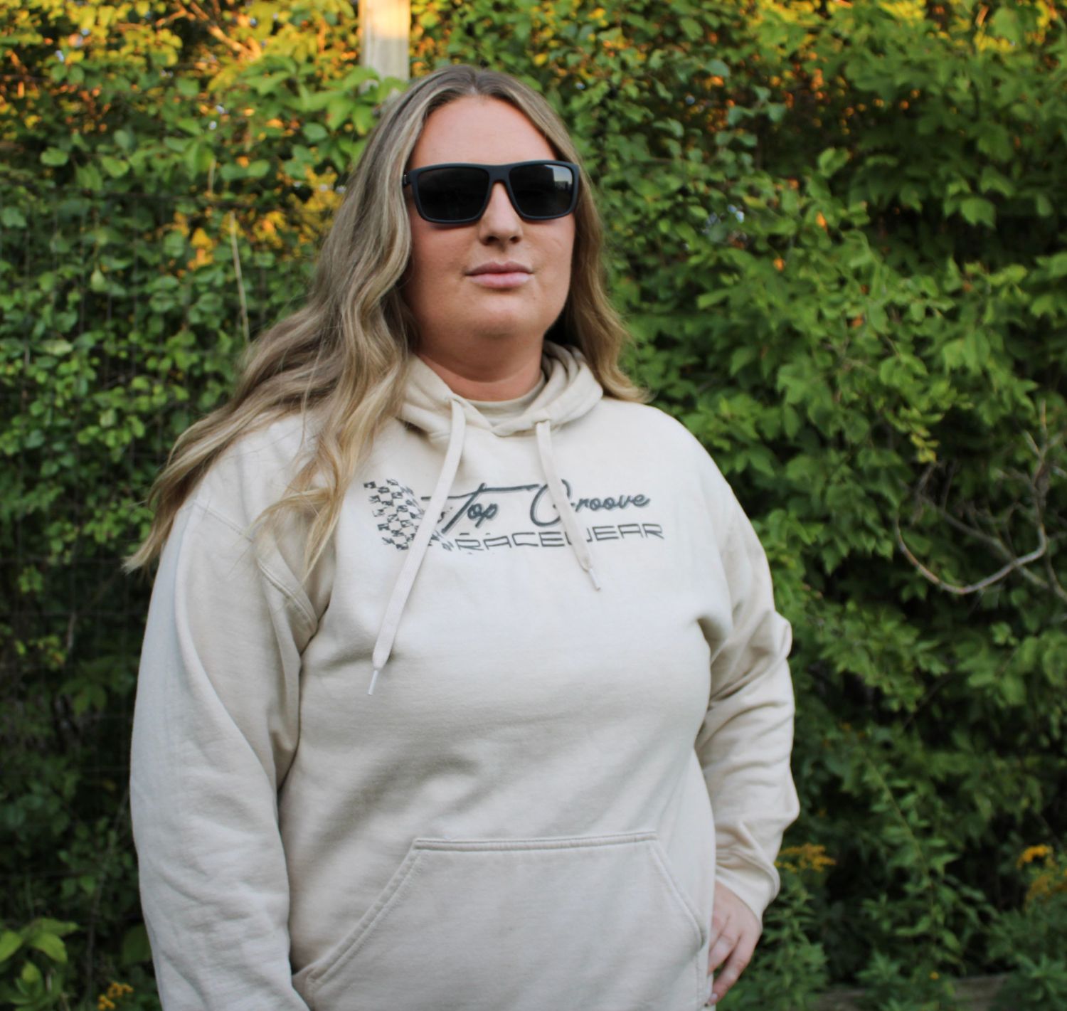 Person modeling a white sweatshirt with custom text design against green foliage.