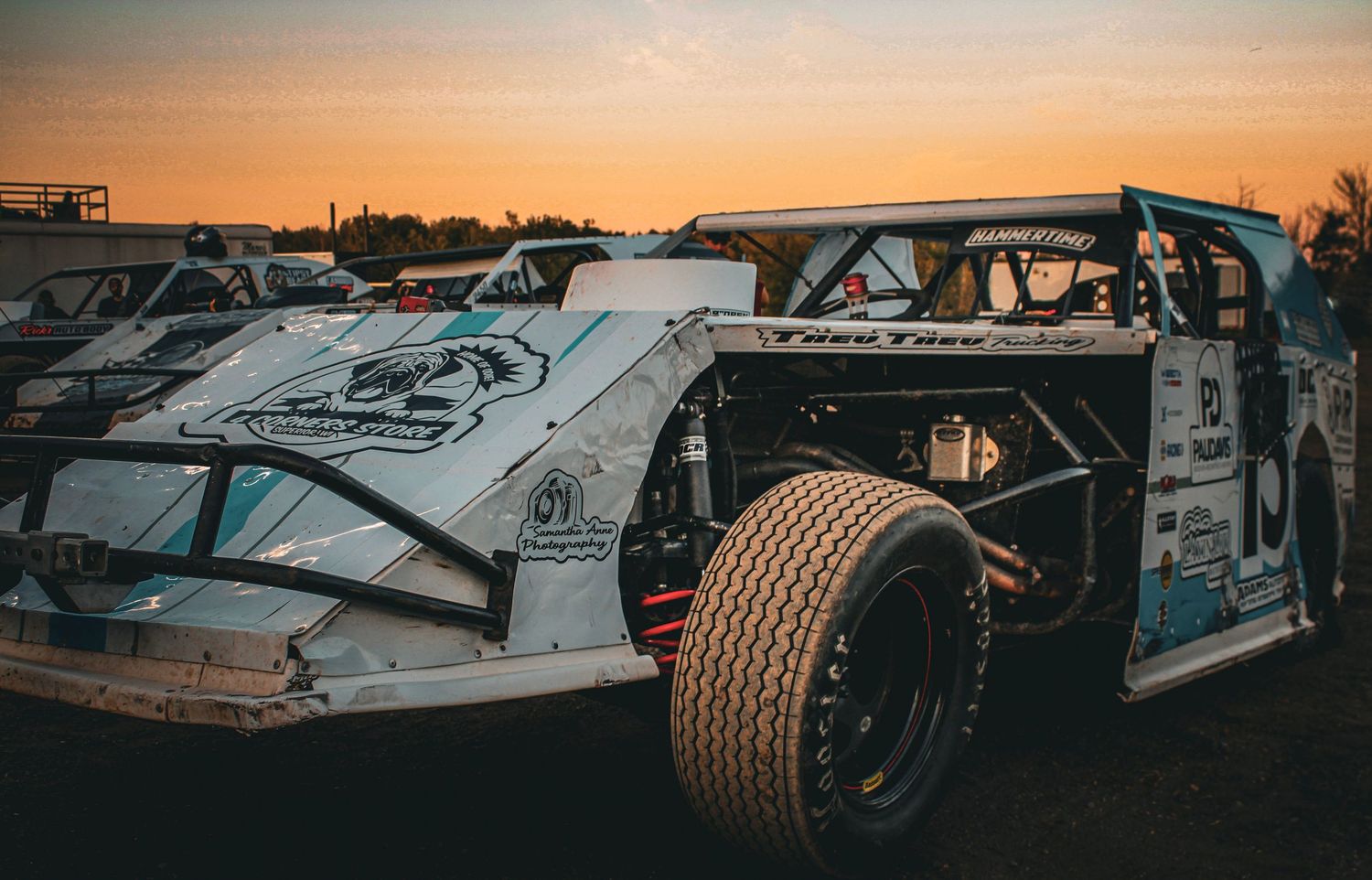 Race cars line up on a dirt track against a dramatic orange sunset sky creating a silhouette effect.