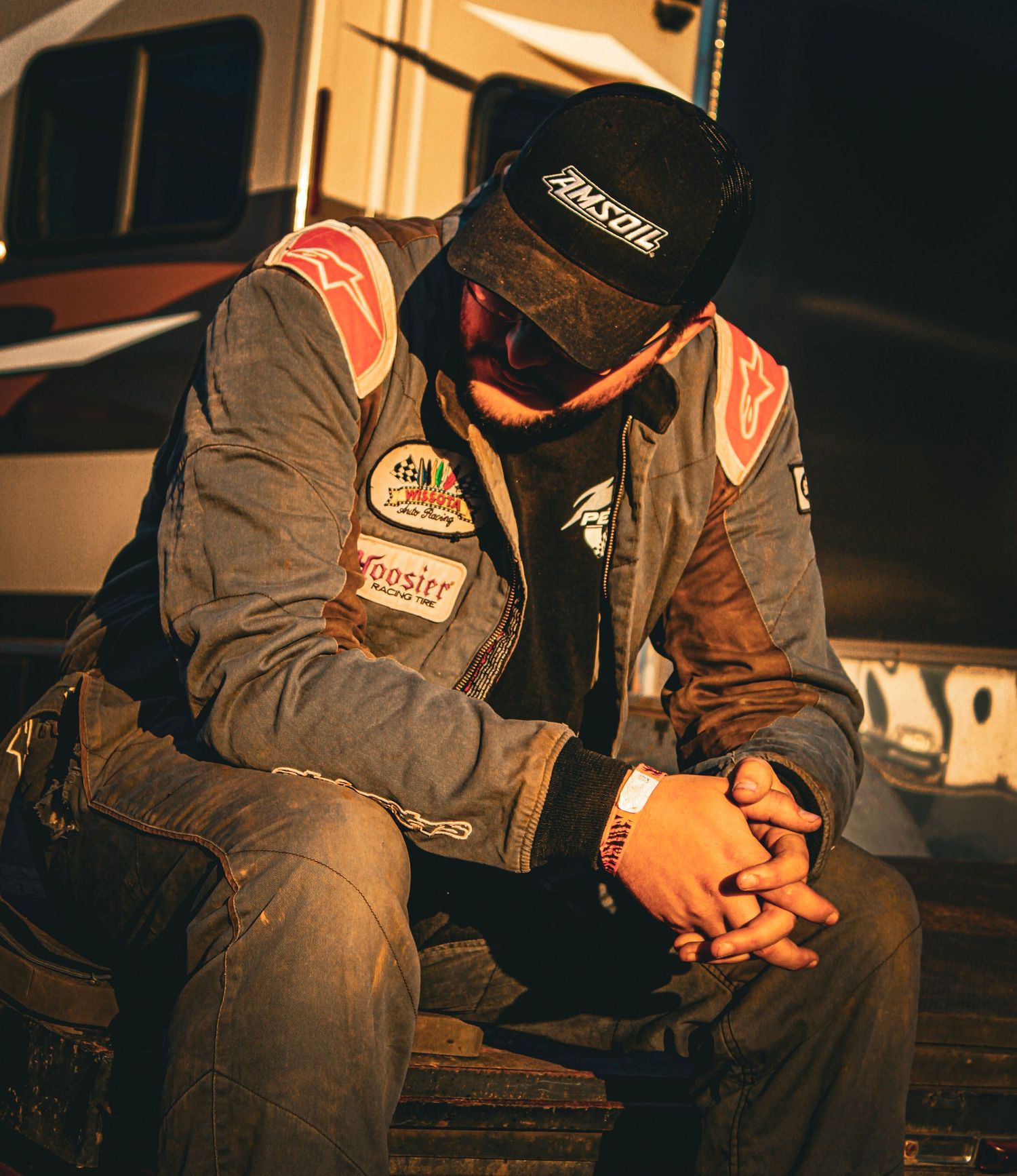A race car driver in a worn racing suit and Amsoil cap sits with head bowed, hands clasped, looking dejected.