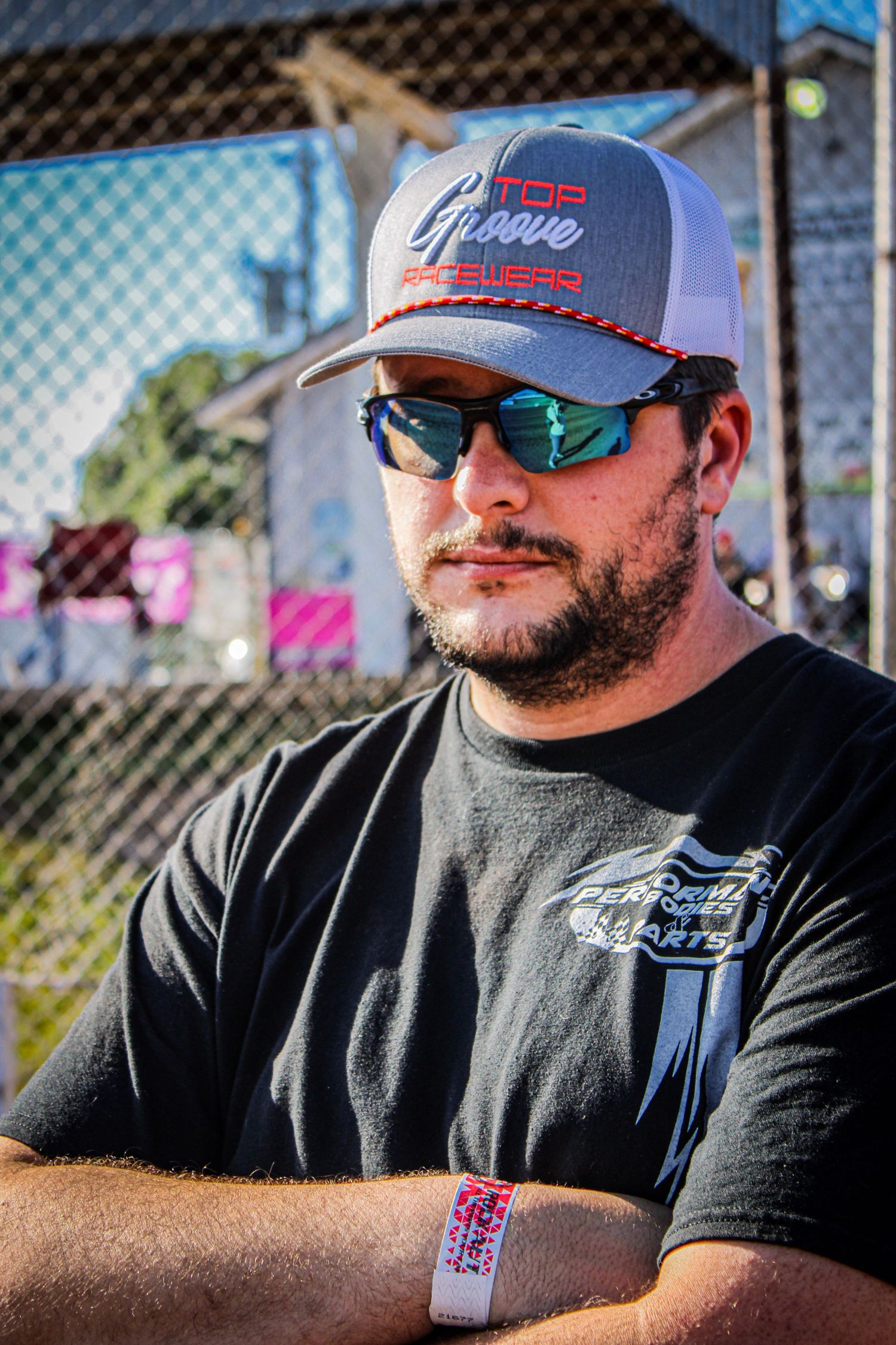 Person wearing sunglasses and baseball cap sitting outdoors on sunny day with fence in background.