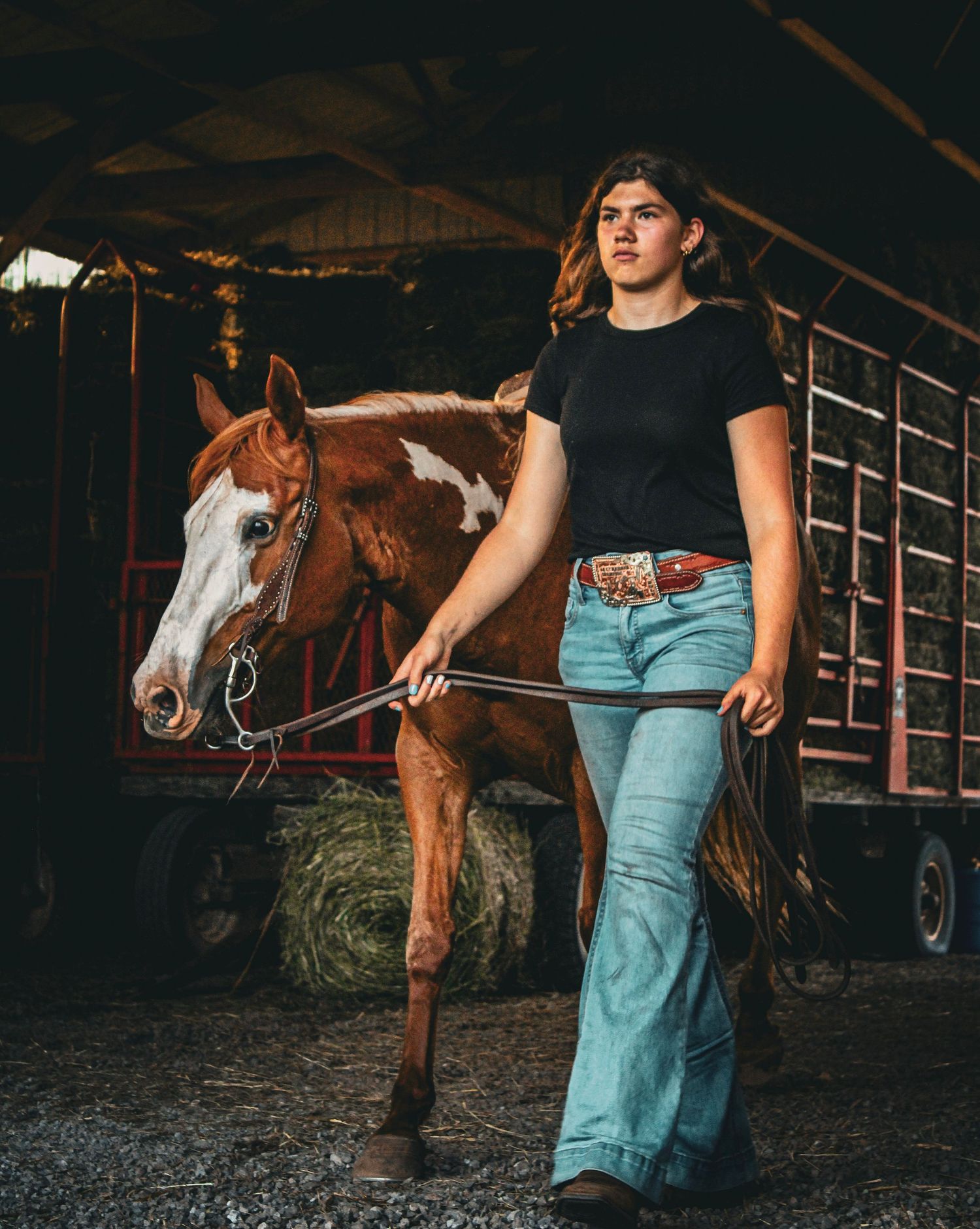 Young woman in jeans and black tee leads a chestnut pinto horse through a rustic barn with hay trailer.