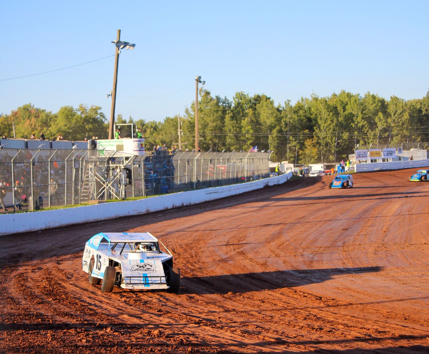 Dirt track racing cars compete on red clay oval speedway surrounded by safety barriers.