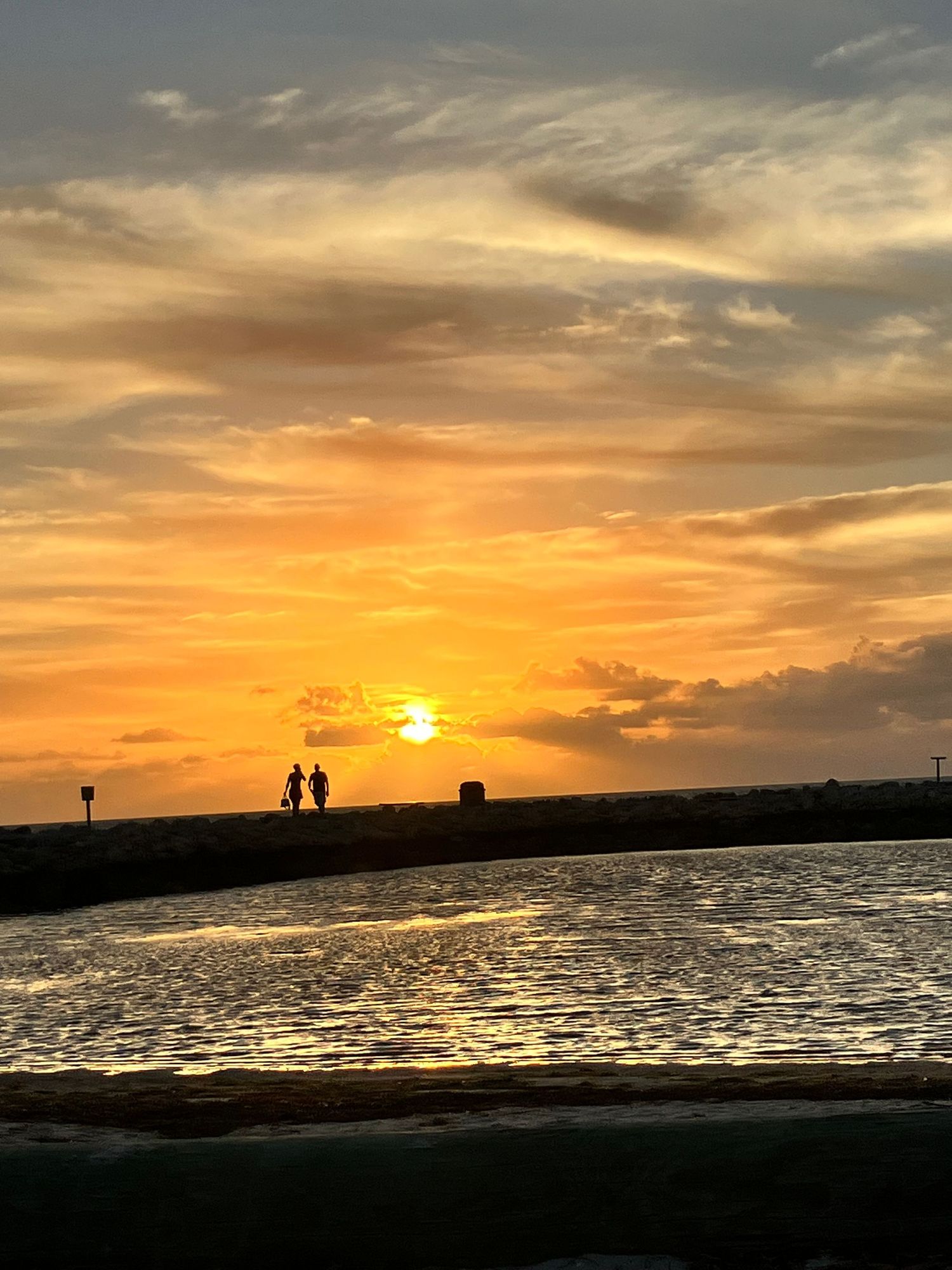 Dramatic orange sunset over water with silhouetted shoreline and cloudy sky.
