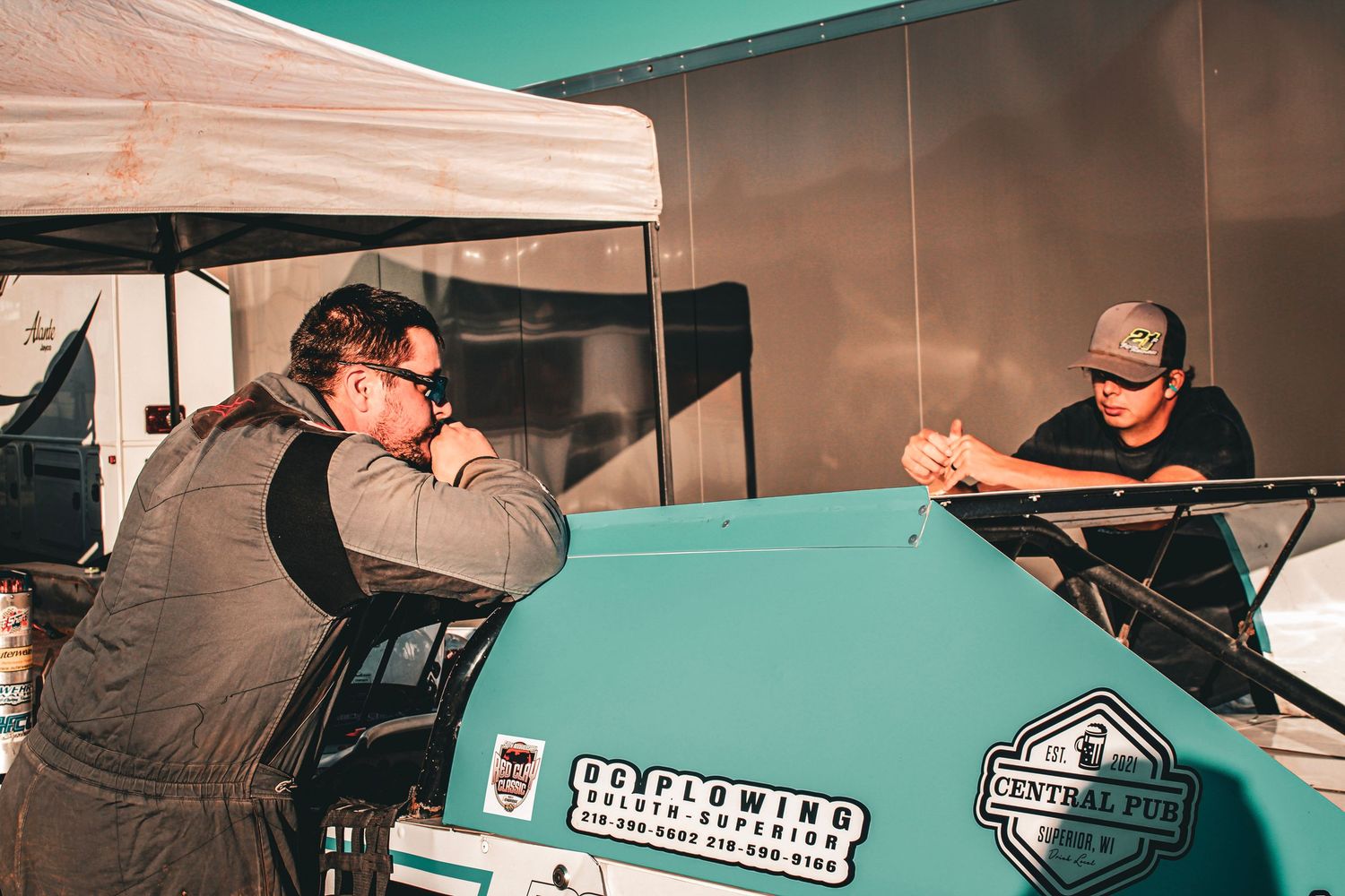 Two crew members discuss strategy while leaning on teal race car under canopy tent.
