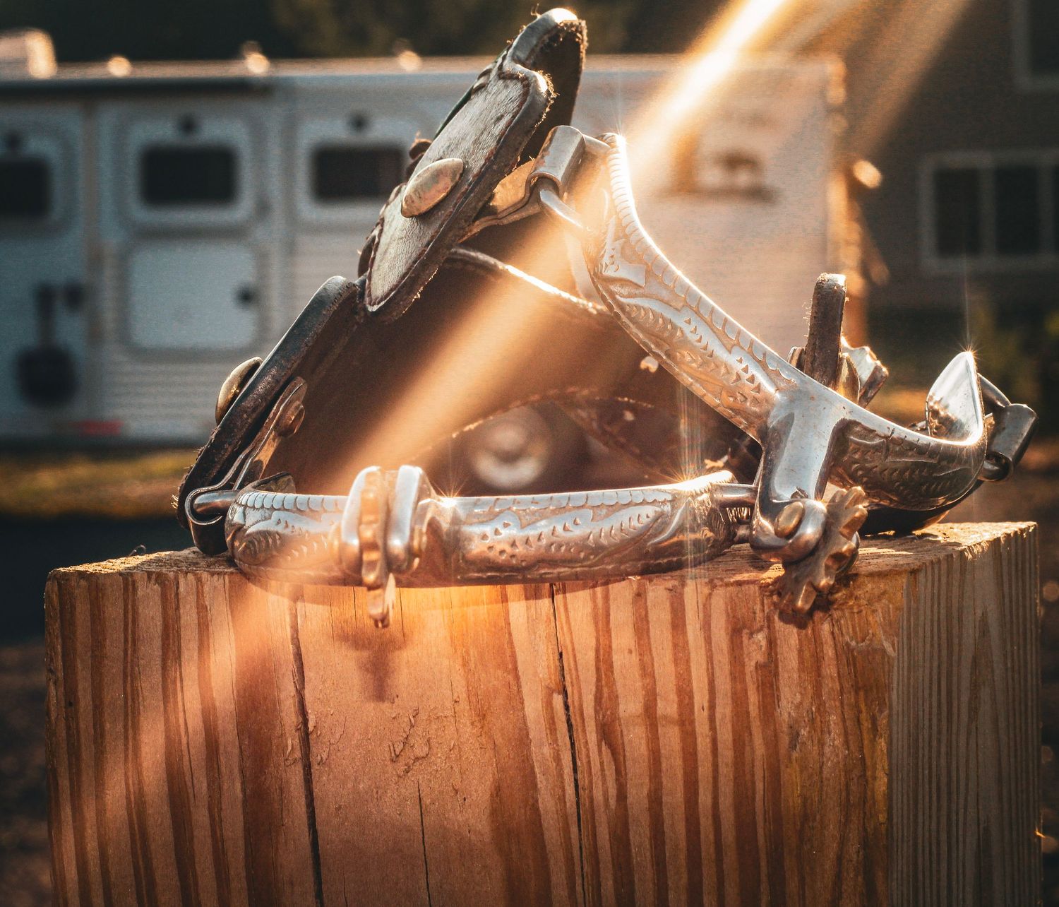 Western spurs resting on a wooden post with golden sunlight rays and a horse trailer in the background.