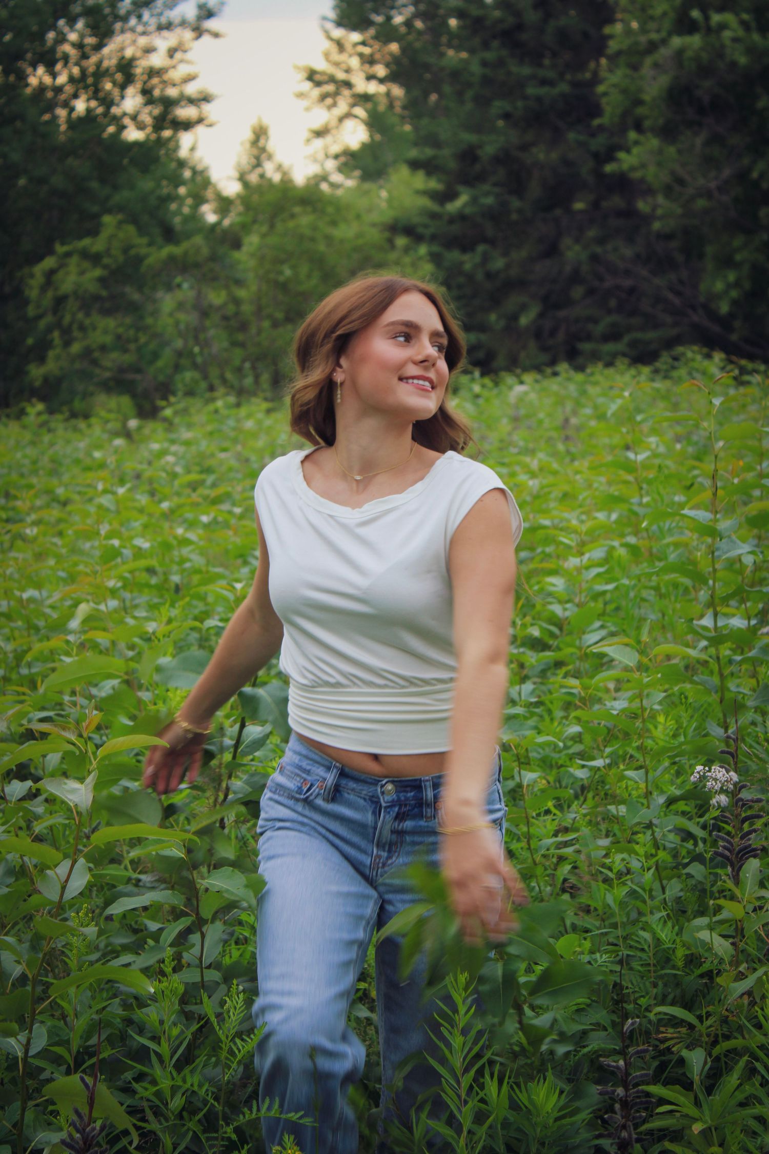 A smiling young woman in a white crop top and jeans walks through a lush green field at dusk.