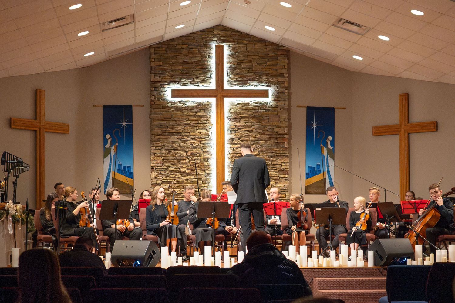 Orchestra performs in church sanctuary with illuminated cross and stone wall backdrop.