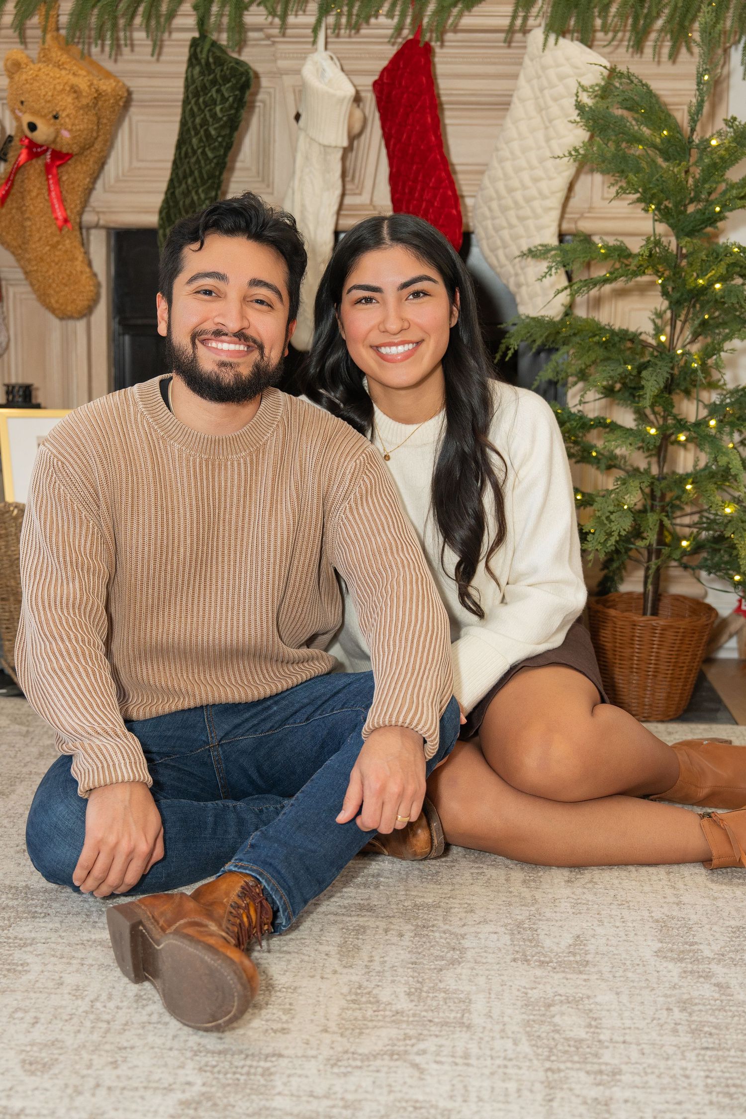 A couple lounges on a white carpet in front of a Christmas tree wearing coordinated sweaters.