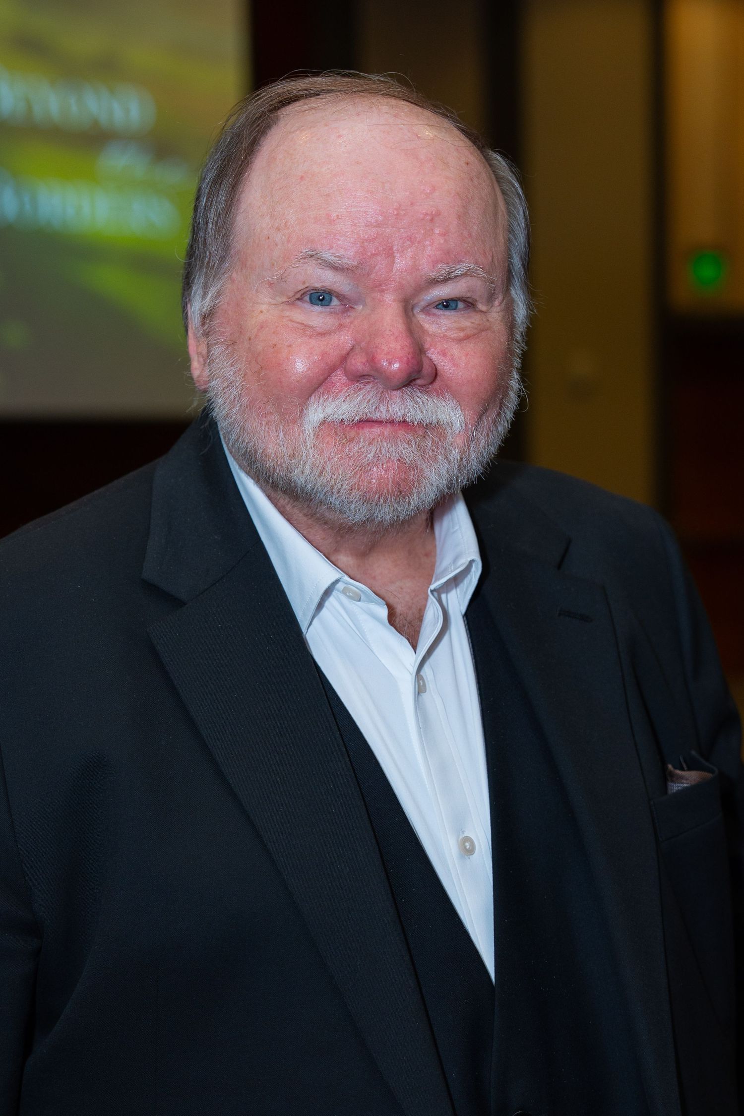 A person wearing a black suit and white shirt smiles warmly at the camera in an indoor setting.