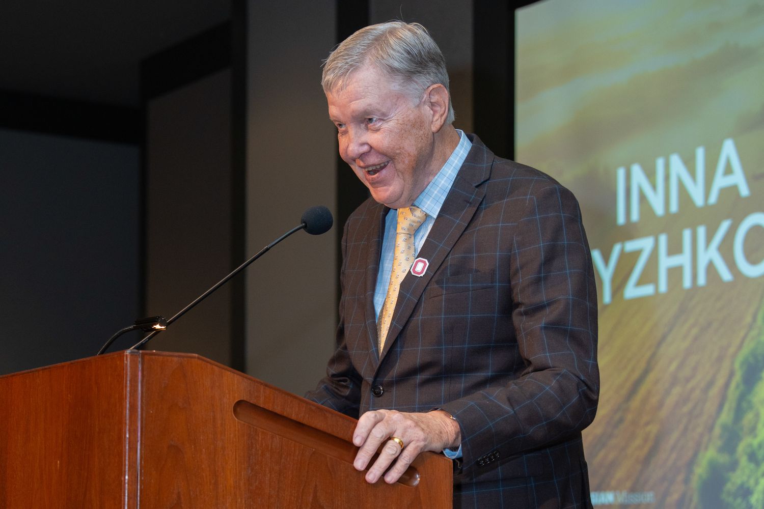 A speaker delivers remarks at a podium with projected presentation visuals in the background.