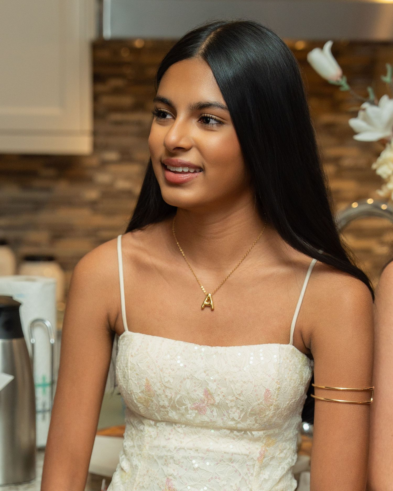 A smiling person in a white lace dress with spaghetti straps and a delicate gold necklace against a blurred background.