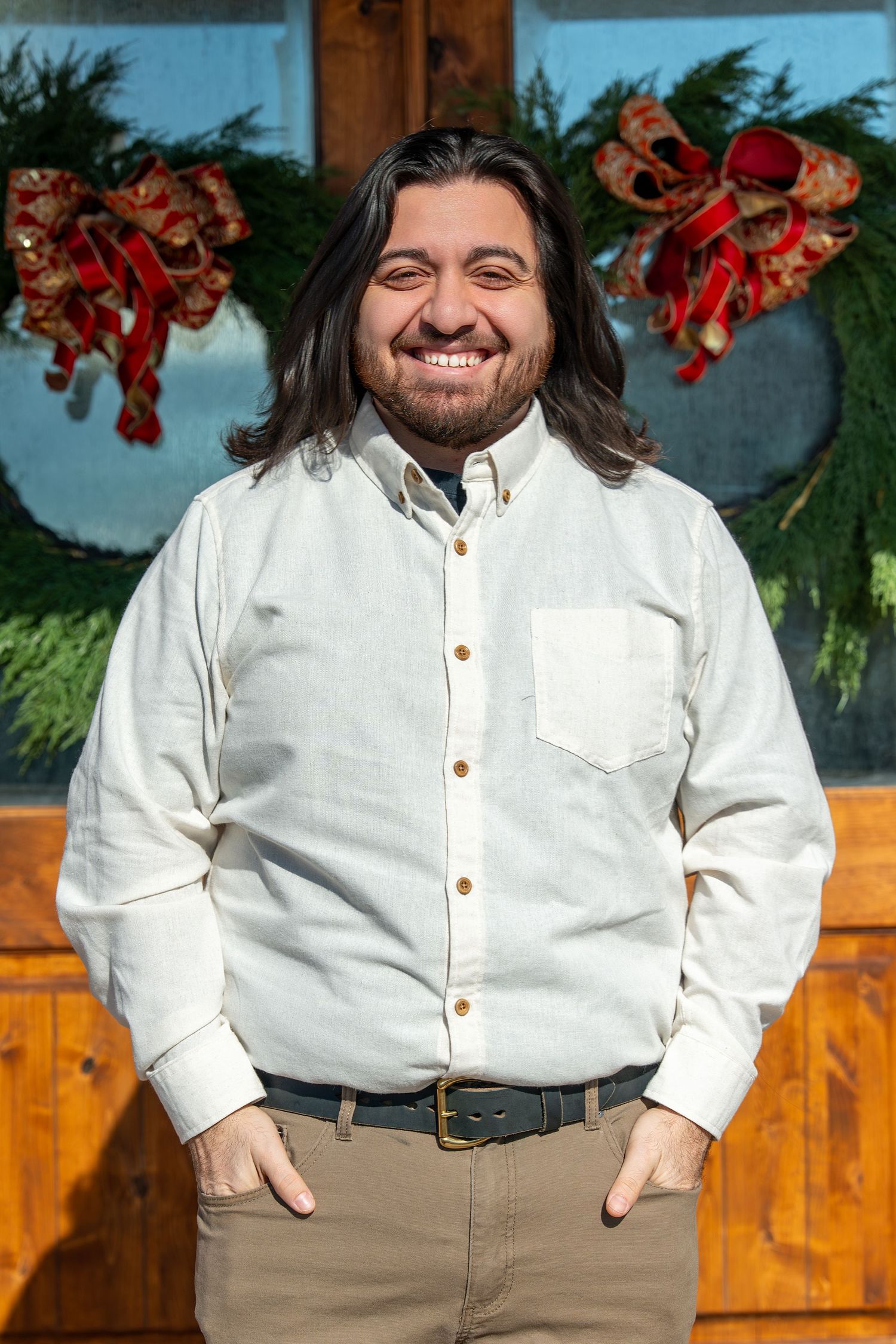 A person wearing a white button-down shirt and khaki pants smiles warmly against a rustic wooden background.