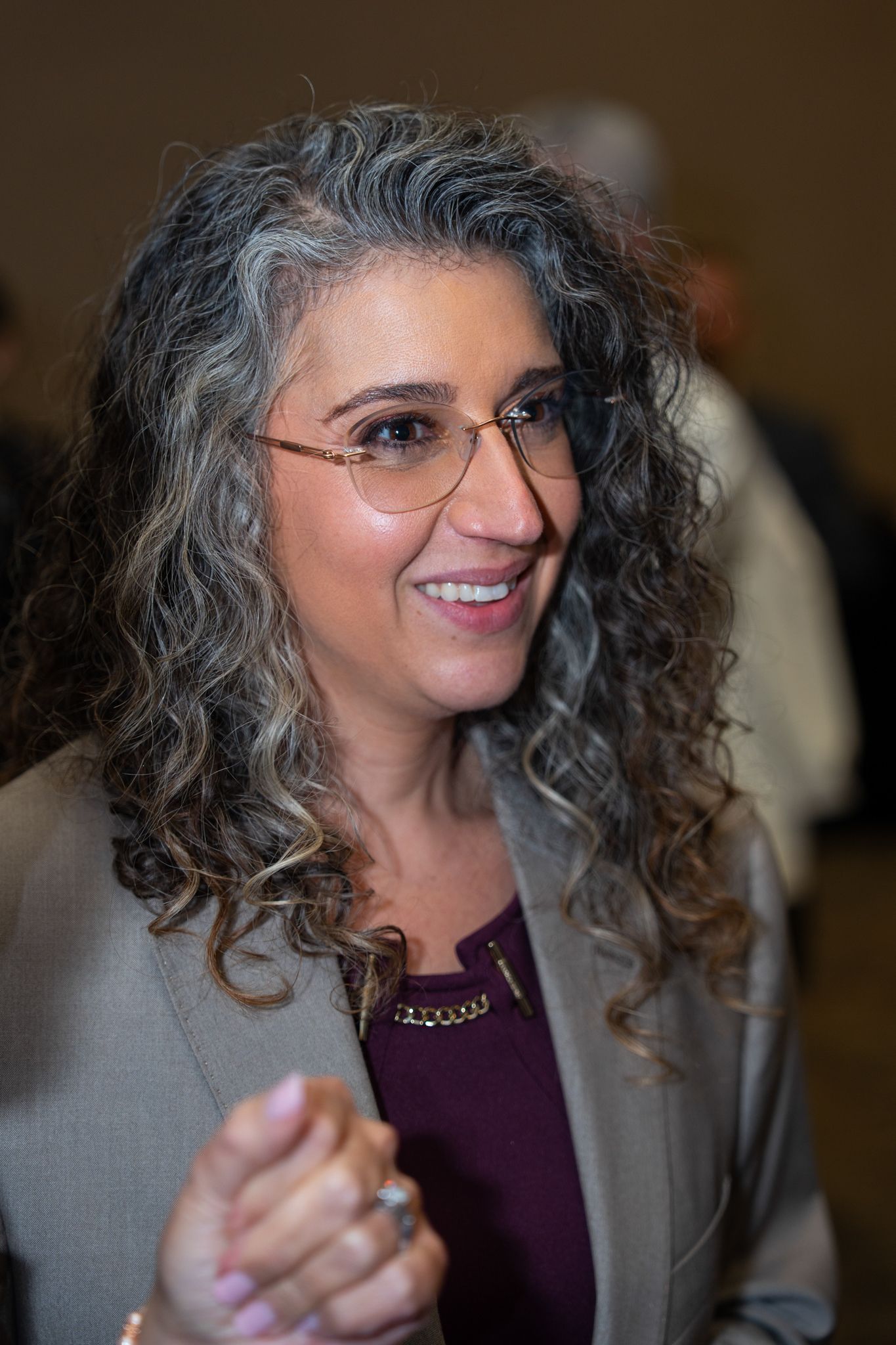 Person with curly gray hair and gray blazer smiling at indoor event.