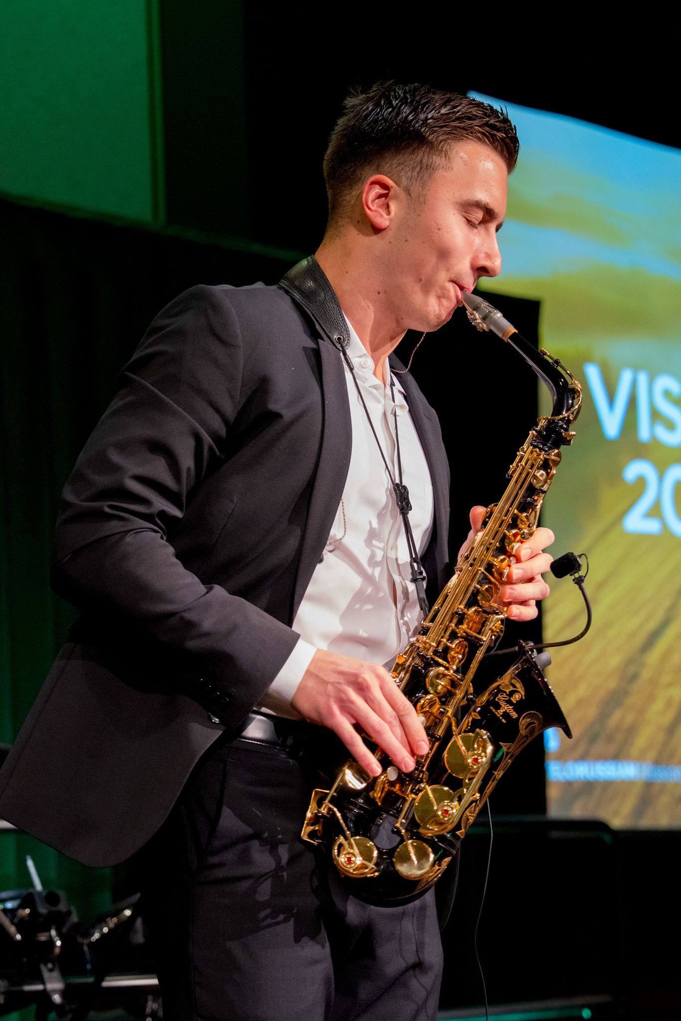 Musician in black suit jacket plays saxophone on stage with projected visuals in background.