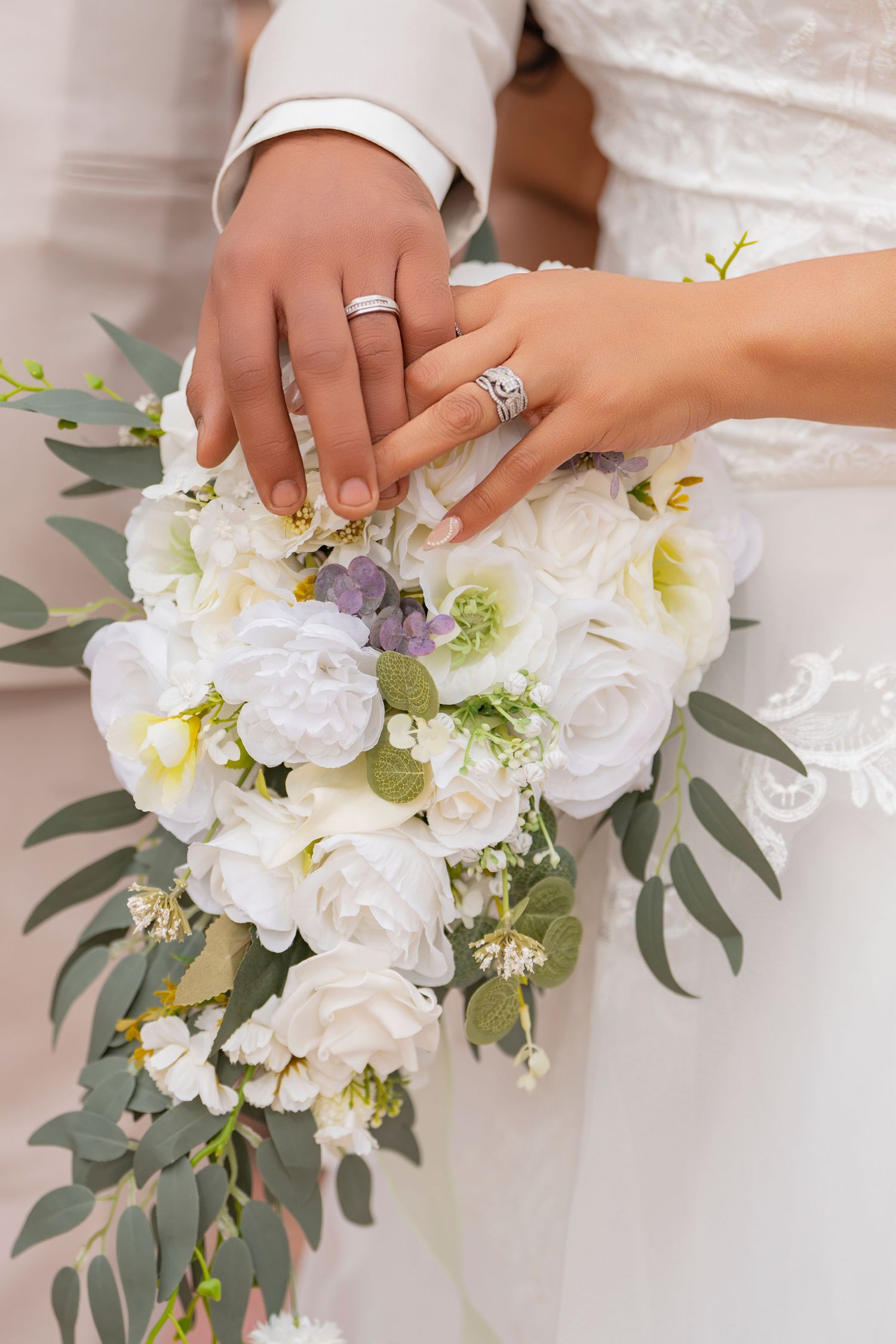 Two hands wearing wedding rings rest on a white and purple floral bouquet with eucalyptus leaves.