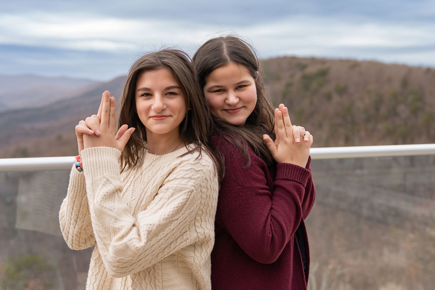 Two people in casual sweaters pose together against a scenic mountain backdrop on a cloudy day.