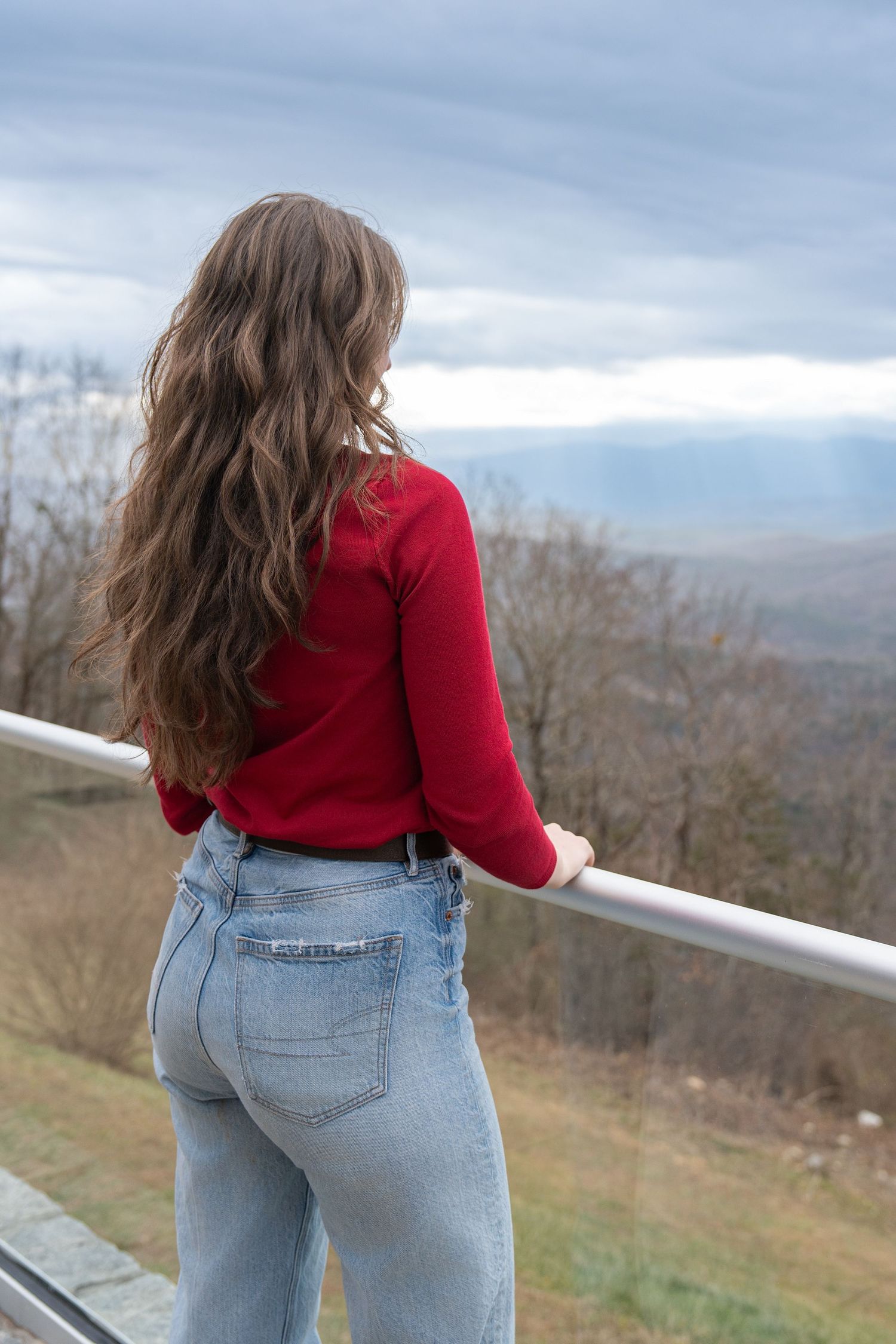 Person in red sweater and jeans looking out over scenic mountain landscape from observation deck.