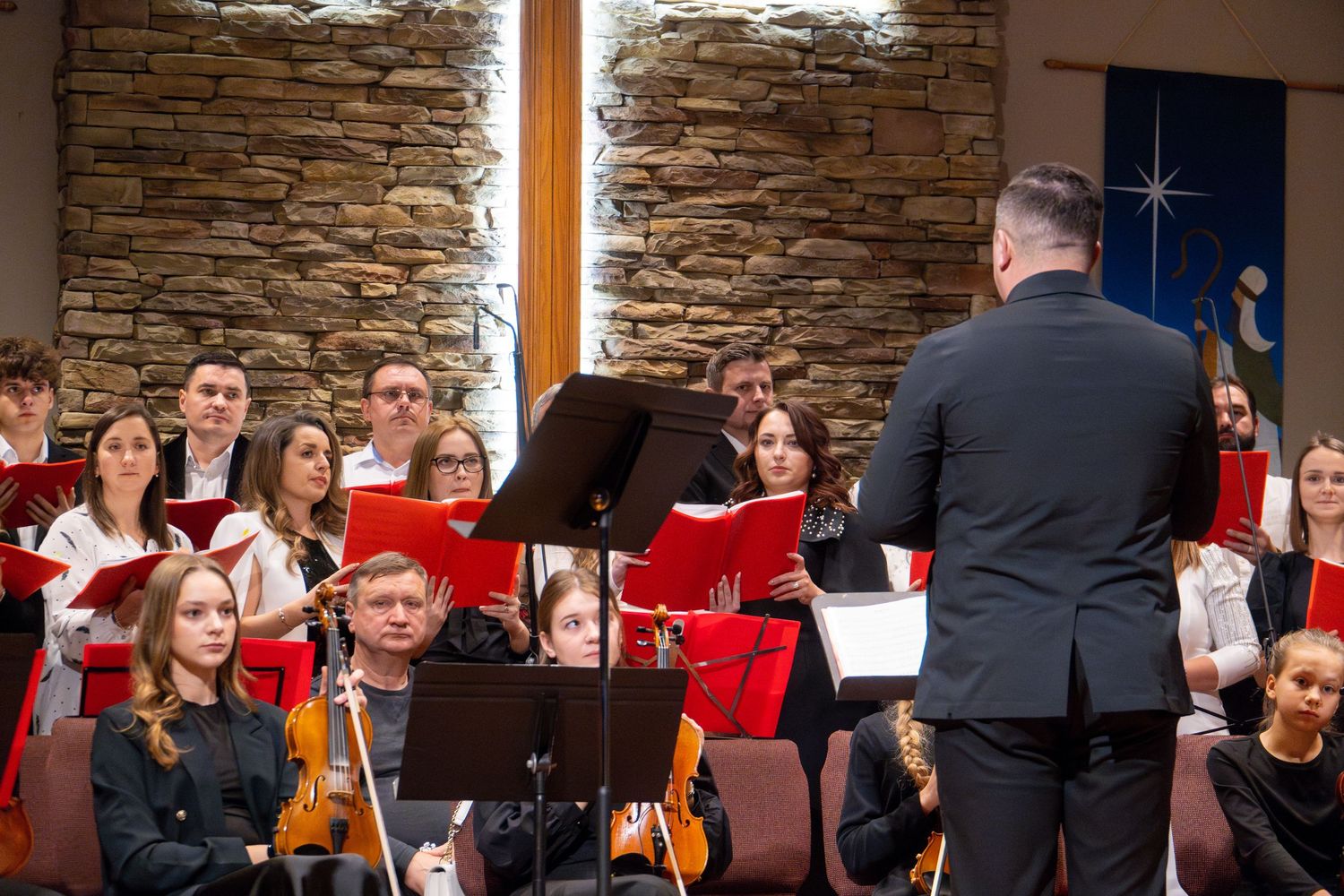 Orchestra conductor leads ensemble in front of stone wall during performance.