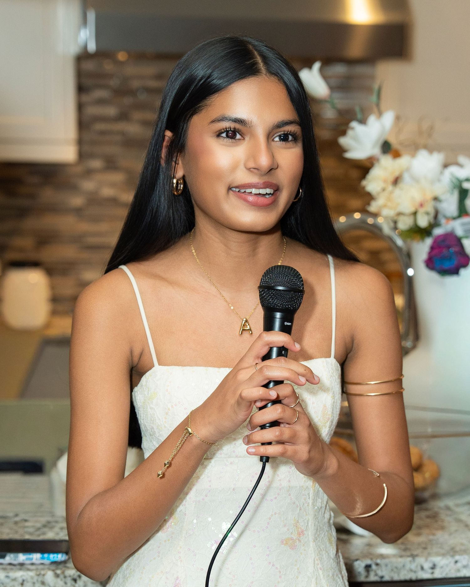 A speaker in a white dress holds a microphone during a special occasion in an elegant indoor venue.