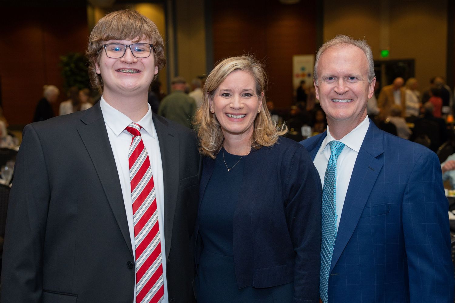 Three people in formal attire pose together at an indoor event, wearing suits and business clothing.