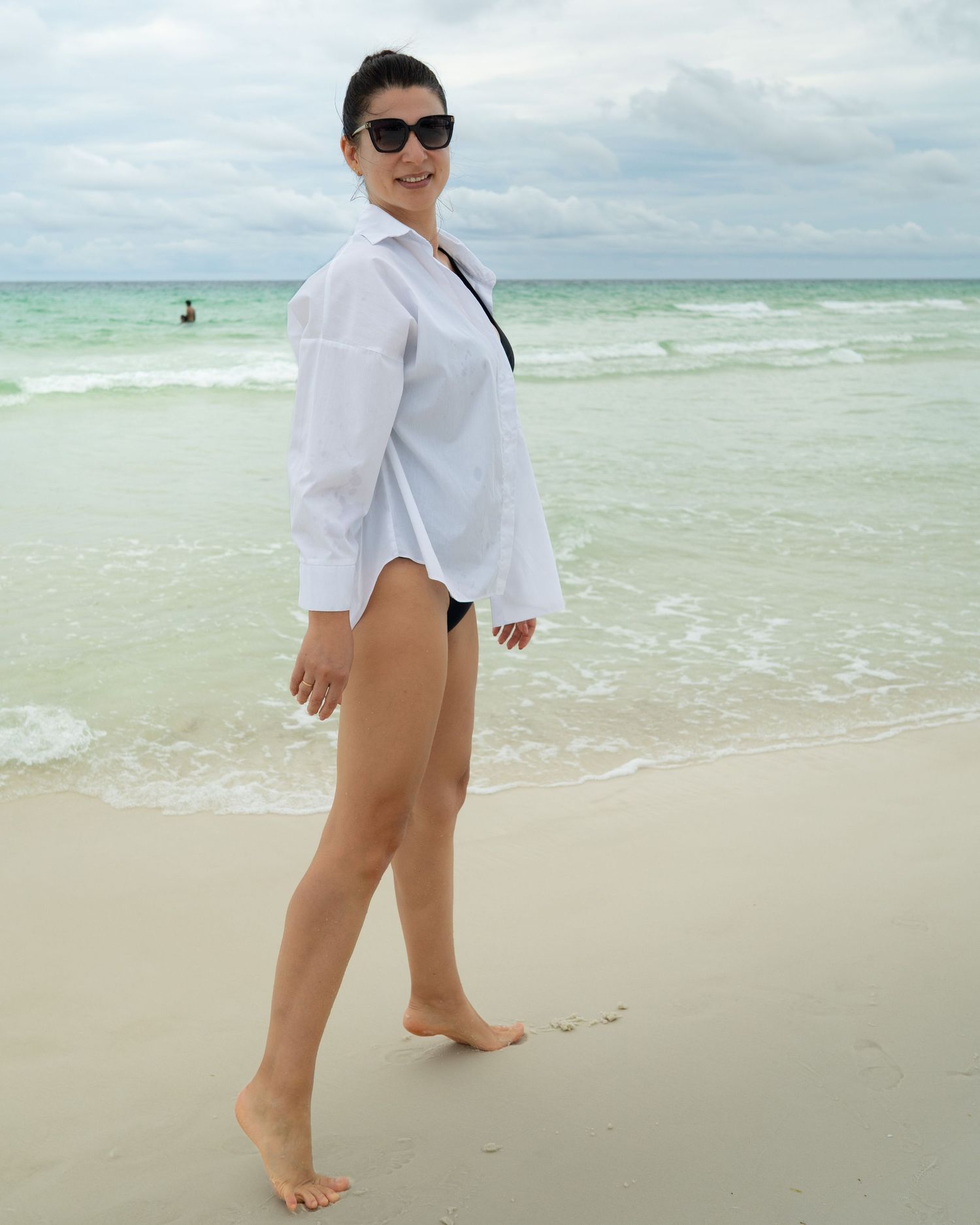 Beach scene with person walking along shoreline wearing white shirt and black swimsuit.