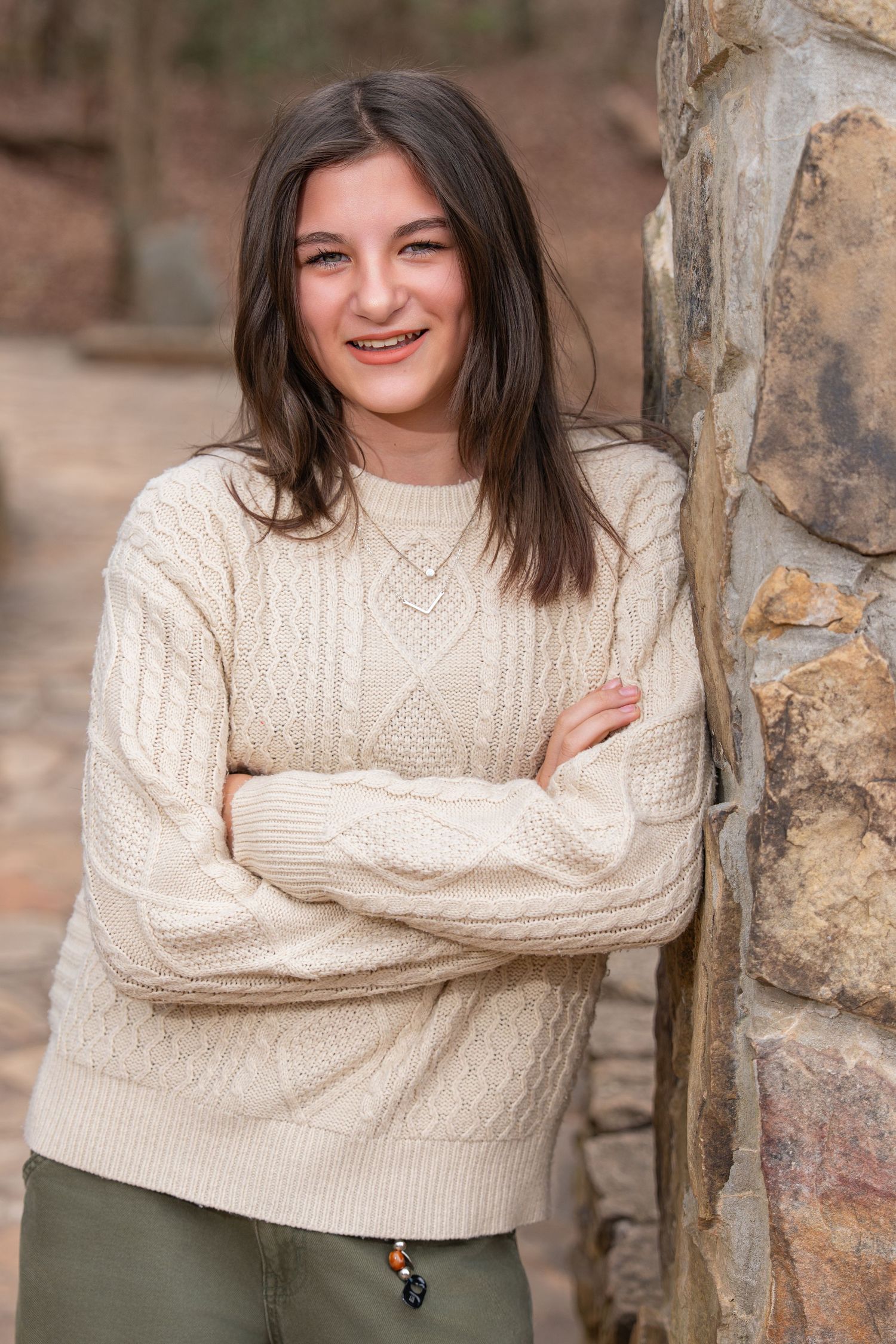 Someone in a cozy cream cable knit sweater and olive pants leans against a stone wall while smiling warmly.