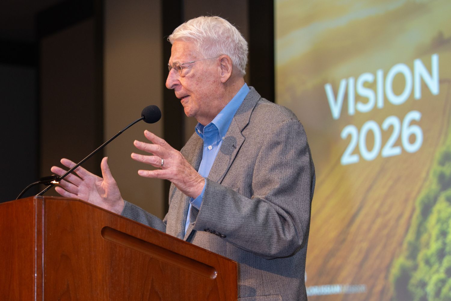 A presenter gestures while speaking at a podium with Vision 2026 displayed on screen.